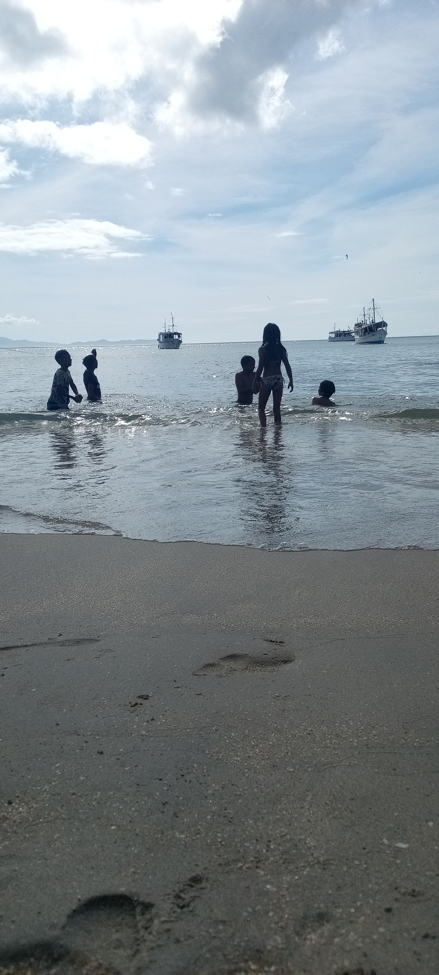 The image shows a group of five children in the ocean near a beach. They are dark-skinned and partially submerged in the water. Behind them, further out in the ocean, are three boats. The sky is a mix of blue and cloudy. The foreground is a sandy beach with footprints. The location is Unknown, Unknown.