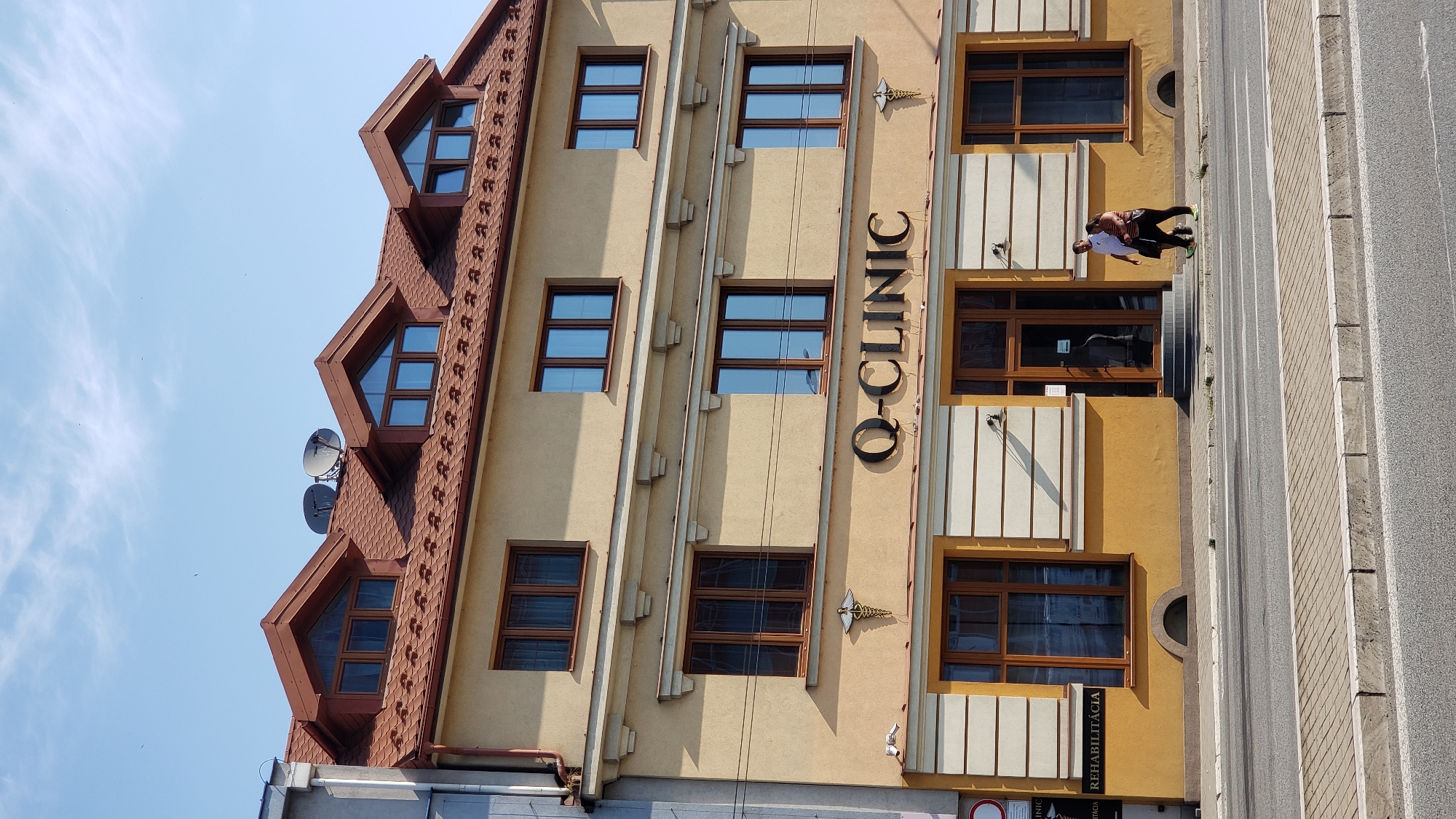 The image shows a three-story building with a yellow facade, a brown tiled roof, and multiple dormer windows. The building's name, "Q-CLINIC," is displayed prominently in large, dark letters above the ground floor entrance. A caduceus symbol, often associated with medicine, is visible to the right of the clinic's name. The ground floor features large windows and a glass entrance door. To the left of the entrance, a sign reads "REHABILITÁCIA." Two individuals are walking on the sidewalk in front of the building. The scene is set on a street with a sidewalk and a road, under a clear blue sky. The building is situated in the city of Prešov, Slovakia.