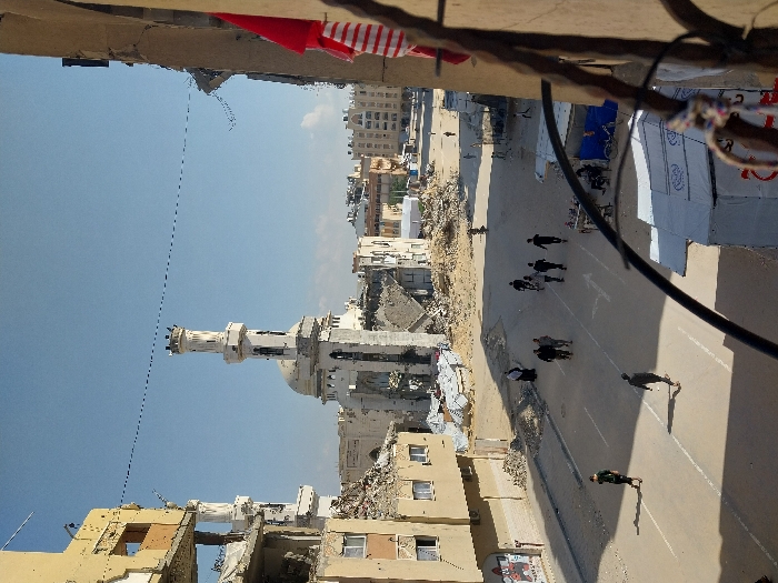 The image displays an elevated, wide-angle view of a street scene in Khan Yunis, Palestinian Territory, under a clear blue sky. The central feature is a large, white mosque with a prominent minaret, both exhibiting substantial structural damage, including partial collapse and visible debris, though their primary forms remain recognizable.
Surrounding the mosque and lining the street are numerous multi-story buildings, primarily light brown or beige, showing extensive destruction. Facades are obliterated, interiors are exposed, and large piles of rubble accumulate at their bases. In the immediate left foreground, a multi-story building is significantly damaged, with visible internal structures. The right foreground includes part of the vantage point, specifically a brown architectural element, a dark metal railing, and hanging red and white striped fabric.
Approximately 15 pedestrians are present on the paved street, which features faded white lane markings and arrows. A group of six individuals walks centrally on the road in the mid-ground. Other people are dispersed, walking alone or in smaller groups. Two white, tent-like temporary structures are visible on the right side of the road, with at least one person seated nearby and a bicycle parked close by. The overall environment is characterized by widespread structural damage, accumulated debris, and a dusty ground surface.