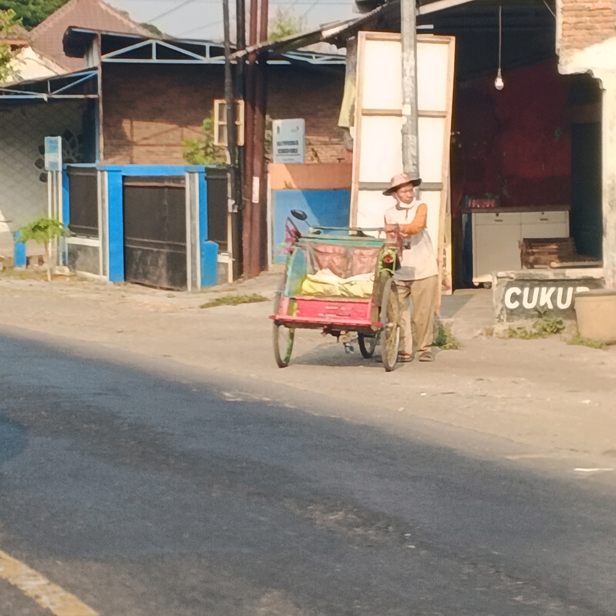 In Menganti, Indonesia, a man in a straw hat and a light-colored shirt with orange accents pushes a three-wheeled pedicab. The pedicab has a red frame and a canvas cover. The man is standing on the right side of the pedicab, looking to his left. The scene takes place on a paved street in front of a building with a corrugated metal roof supported by white metal beams. The building has brown brick walls and a blue and black facade. A sign with "CUKU" is visible on the wall to the right of the building. The ground is a mix of asphalt and dirt. The sun is casting shadows, suggesting it is daytime.