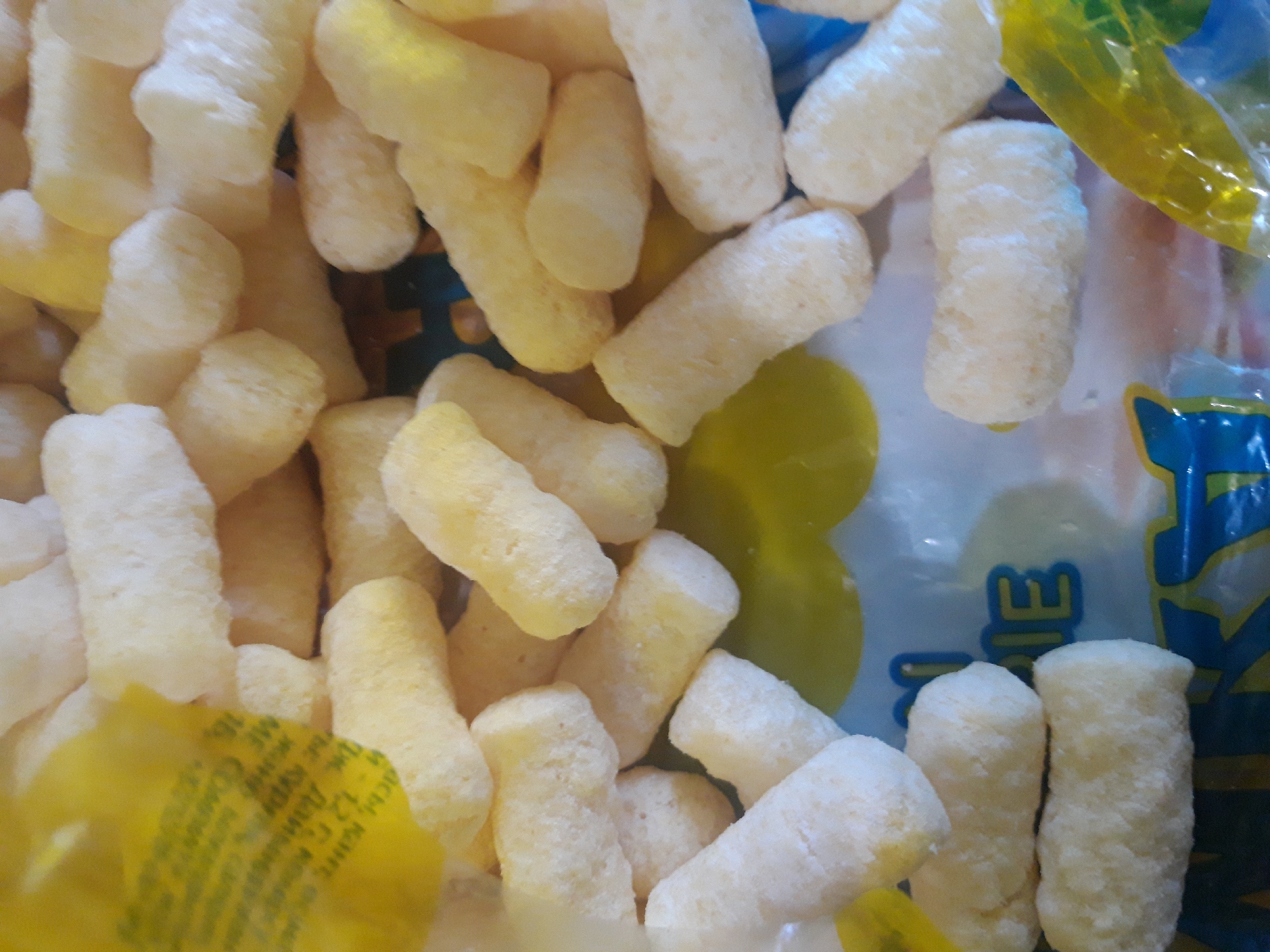 The image shows a close-up of puffed corn snacks. The snacks are cylindrical and light yellow in color, with a slightly textured surface. They are piled loosely in a transparent plastic bag, which has some yellow and blue printed text and graphics visible on it. The lighting is bright and direct, creating some glare and shadows. The background is out of focus but appears to be a flat surface. There are no people or animals present in the image. This image is not related to the city of Макаров ауылдық округі, Kazakhstan.