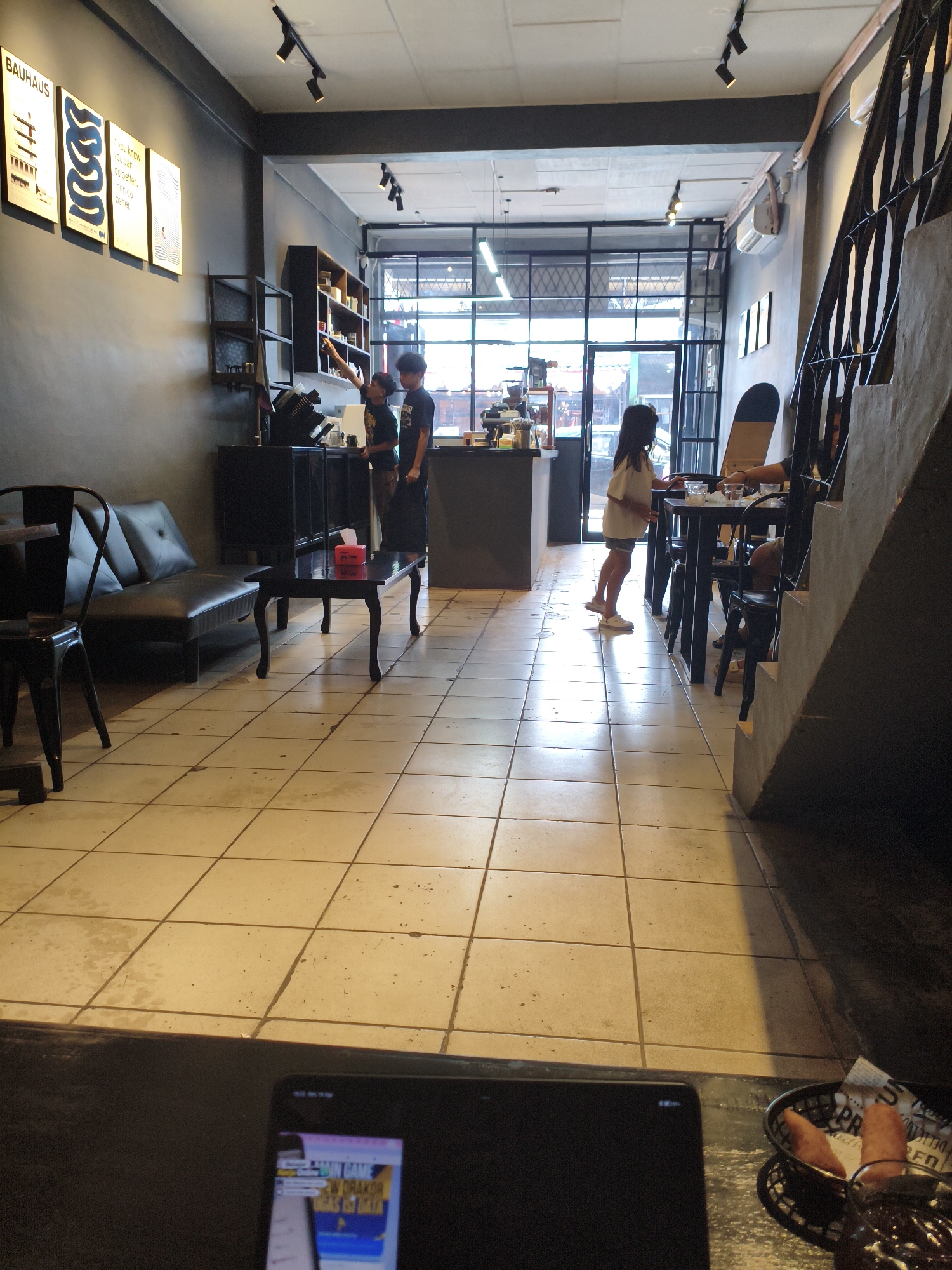 The image depicts the interior of a cafe with a modern industrial aesthetic. The walls are dark grey, and the floor is tiled with large, light-colored square tiles. A staircase with a metal railing is visible on the right side of the frame.
In the foreground, a black leather sofa and two black metal chairs are arranged, suggesting a seating area. A small black coffee table with a red tissue box is placed in front of the sofa.
Further into the cafe, a counter area is visible with two people behind it, appearing to be staff. One is wearing a dark shirt and the other a green shirt. They are interacting with a customer, a young girl in a white t-shirt and denim shorts, who is standing near the counter. Another customer is seated at a table on the right, partially obscured by the staircase.
On the left wall, several framed posters are displayed, including one with the word "BAUHAUS" prominently featured. Shelving units filled with items are also visible against the back wall. The cafe appears to have large windows at the back, allowing natural light to enter. The overall impression is a clean and contemporary space. The scene is likely in a city, possibly Tebing Tinggi, Indonesia, given the context of the request, with a focus on creating a comfortable and stylish atmosphere for patrons.