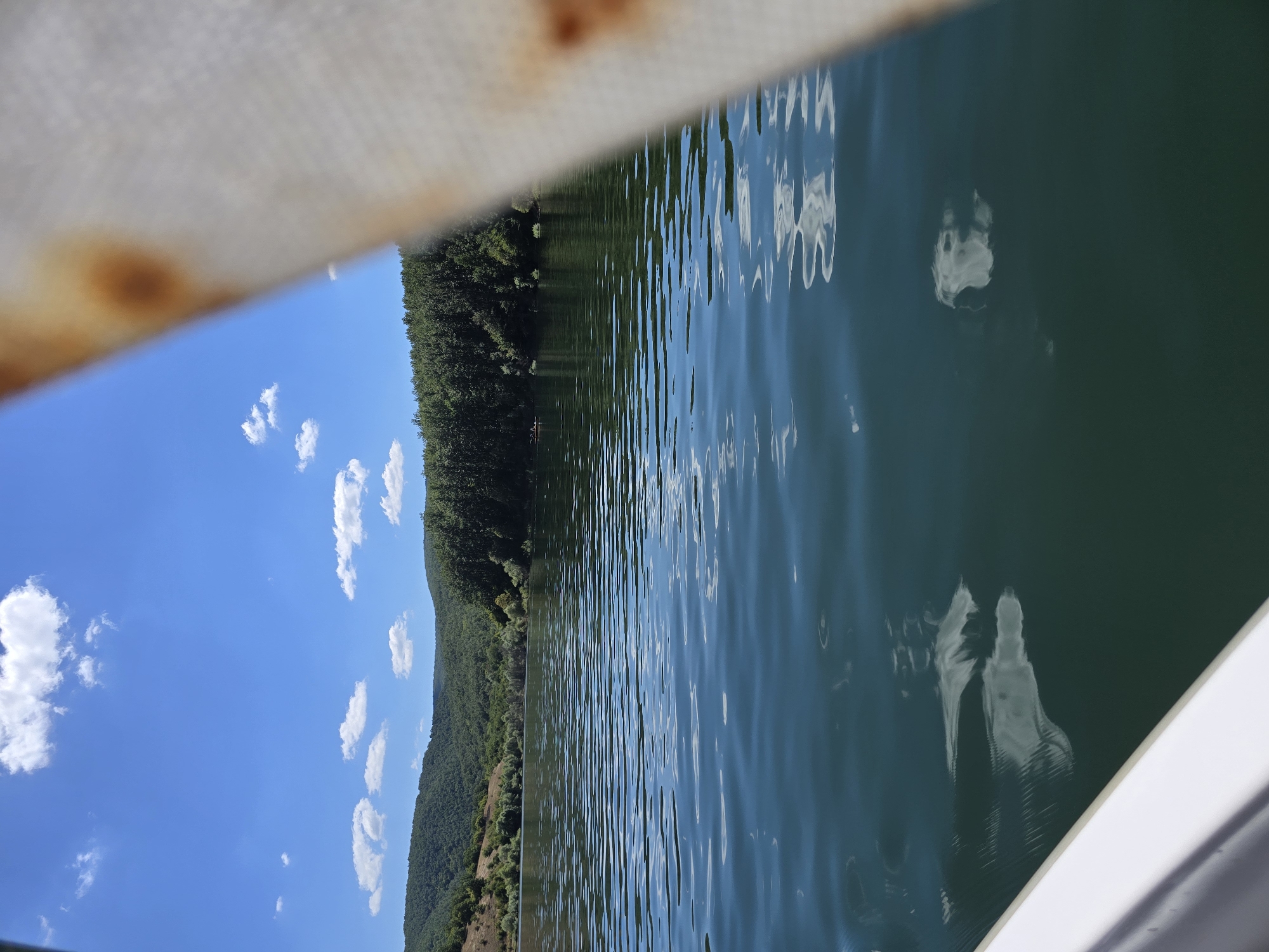A body of water reflects the blue sky and white clouds, with green, tree-covered hills in the background. A white railing is visible in the foreground, partially obscuring the view. The water is calm with gentle ripples. The sky is clear blue with scattered white clouds. The hills are densely wooded with a mix of deciduous and evergreen trees.