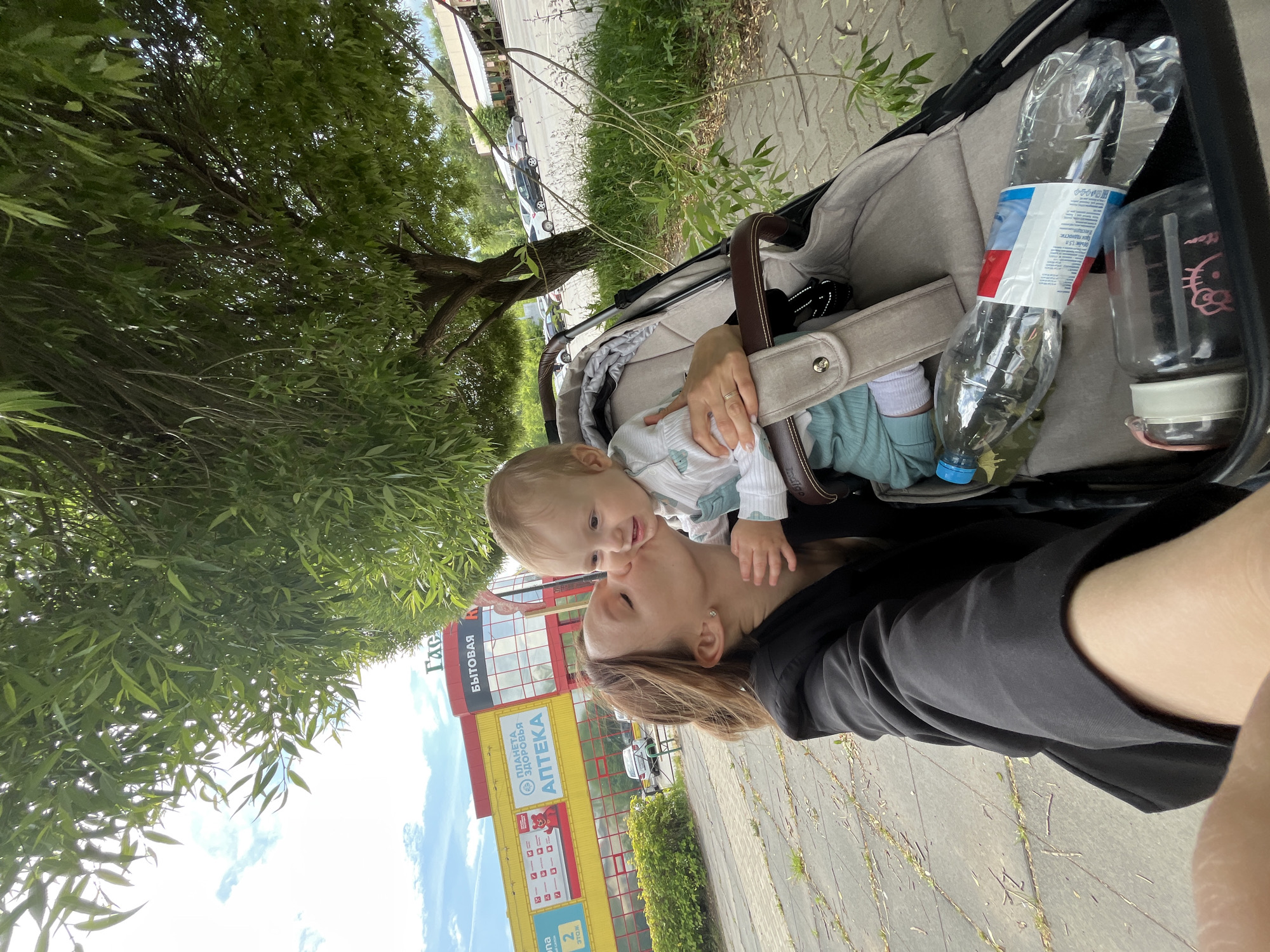 A medium shot photograph captures a woman kissing a baby while standing next to a stroller under a tree in Chaikovsky, Russia. The woman is wearing a black blazer and has her hair pulled back. The baby is sitting in the stroller, wearing a light blue outfit and a white top with a patterned design. A large plastic water bottle and a pink-capped thermos are in the stroller. In the background, a bright yellow and red building with signs in Russian, including "Планета Аптека" (Planet Pharmacy), is visible. A paved walkway and grassy area with trees are also part of the scene. The sky is cloudy with patches of blue.