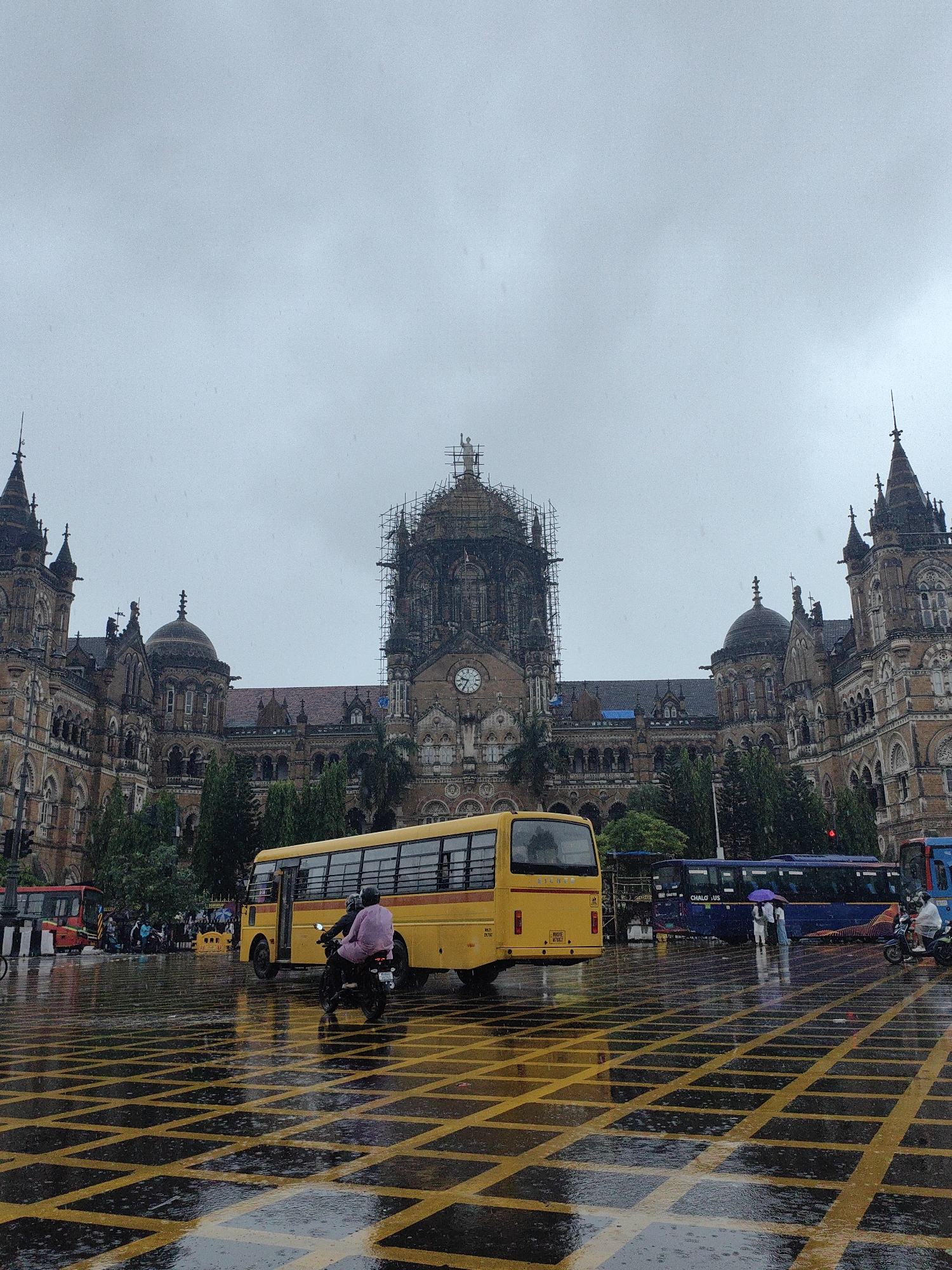 The image depicts a rainy day outside the Chhatrapati Shivaji Maharaj Terminus (CSMT) in Mumbai, India. The grand Victorian Gothic and Art Deco architecture of the building is visible in the background, with scaffolding on one of its domes suggesting ongoing renovation work. The foreground shows a wet street with yellow checkered markings reflecting the overcast sky and traffic lights. A yellow school bus is parked on the right, with a person on a motorcycle in front of it. Further down the street, a red bus and a blue bus are also visible, along with a few pedestrians carrying umbrellas. The scene is typical of Mumbai during the monsoon season, with heavy rain affecting visibility and traffic.