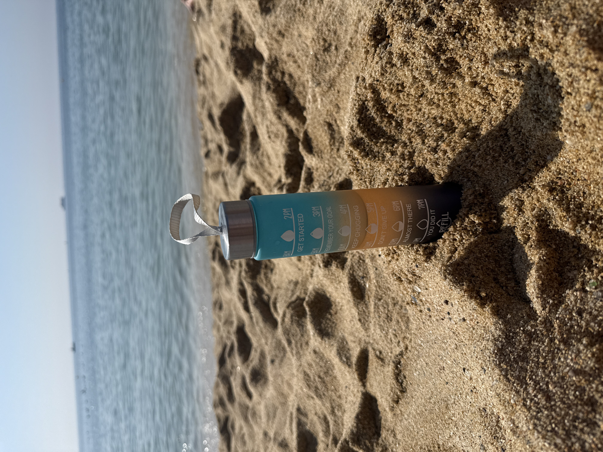 A water bottle with time markings sits on a sandy beach in front of the ocean. The bottle is teal and orange and has markings for 8 AM, 10 AM, 12 PM, 2 PM, 4 PM, 6 PM, and 7 PM. The 7 PM marking says "YOU DID IT REFILL". The water is calm and there is a faint hazy horizon with a distant ship. The sand is light brown and shows ripples from the wind. The image appears to be taken during the day with bright sunlight.