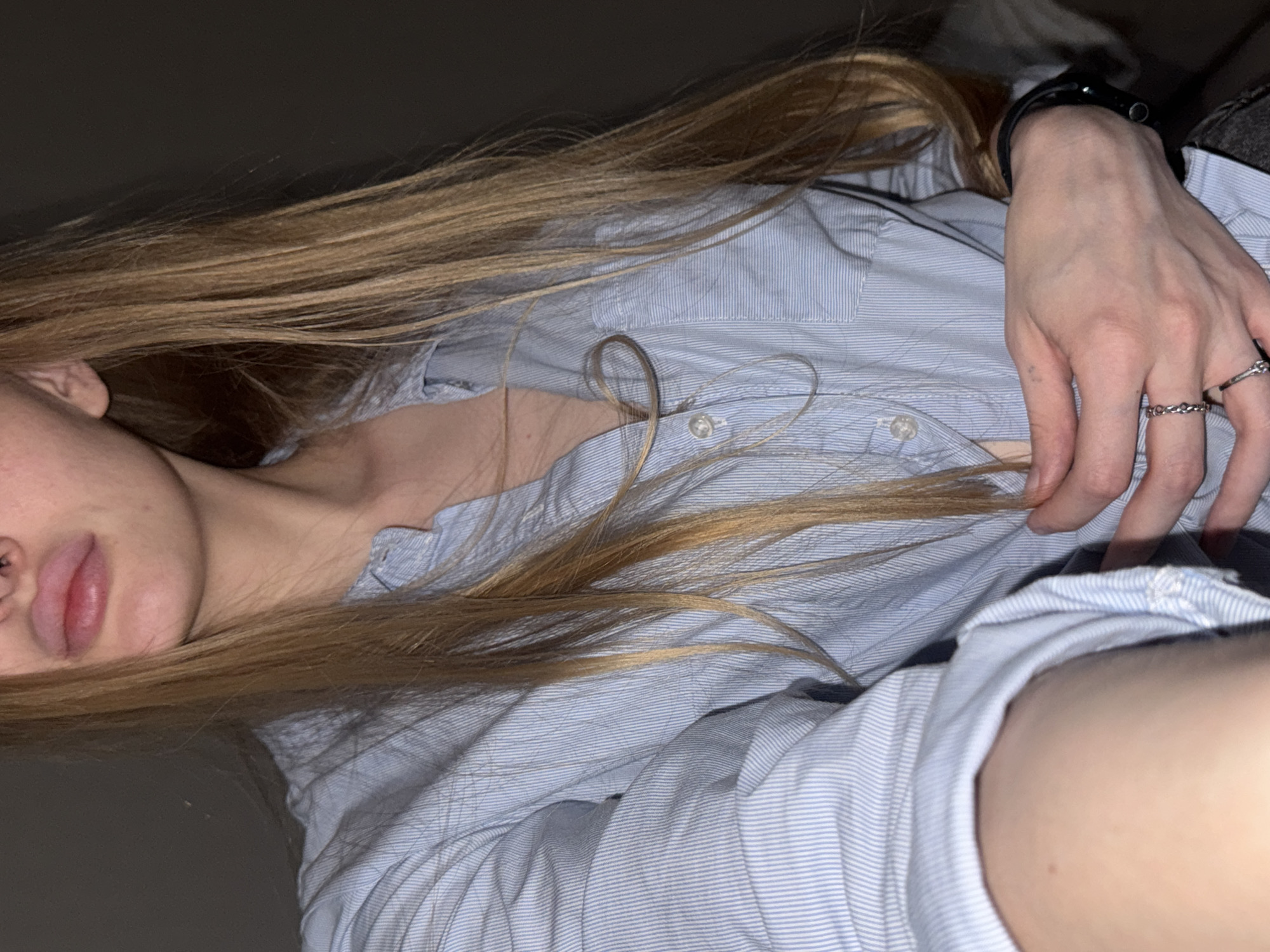 A medium shot shows a person from the chest up. The person has long, blonde hair that falls over a light blue, pinstriped button-up shirt. The shirt is unbuttoned, revealing a portion of the chest. The person's hands are visible, one holding the shirt and the other resting on the chest. The person is wearing rings on their fingers and a watch on their wrist. The background is dark and out of focus. No specific location in Perm, Russia, can be determined from this image.