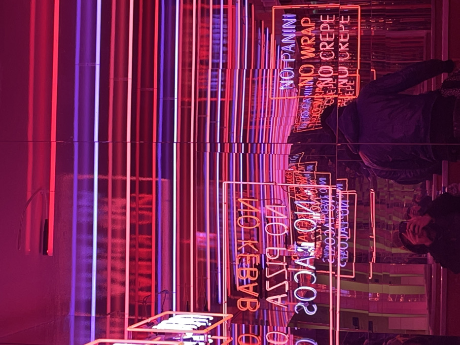 Red and purple neon signs displaying "NO KEBAB," "NO PIZZA," "NO TACOS," "NO PANINI," "NO WRAP," and "NO CREPE" are mirrored in a reflective surface. Straight vertical lines of the same colors add to the distorted and repetitive nature of the image. People are visible through the reflection, adding a human element to the urban, neon-lit scene. The overall effect is surreal and visually engaging.