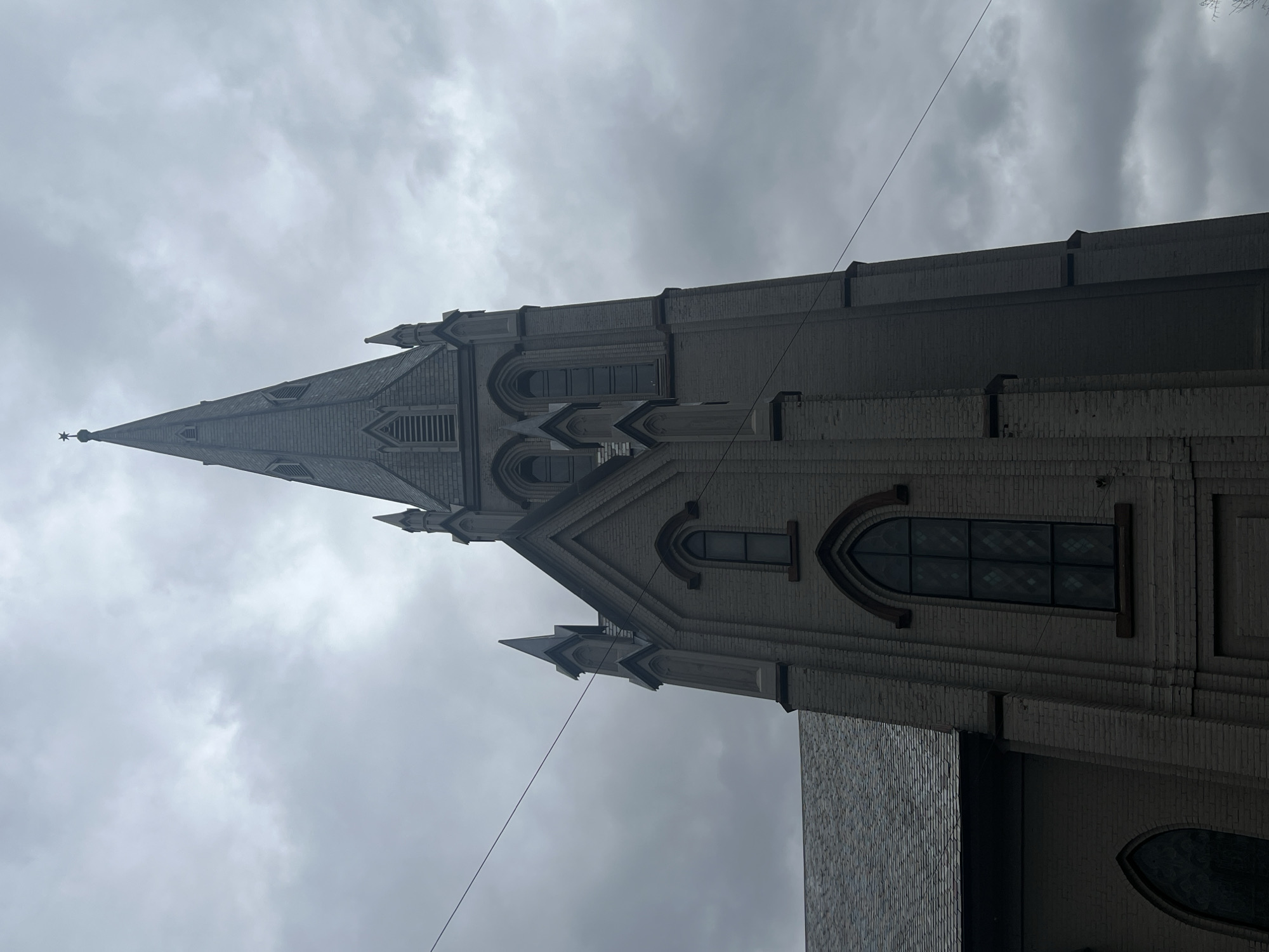 The image captures the exterior of a church in the City of Newburgh, United States of America. The structure is predominantly constructed of light-colored brick and features a tall, pointed steeple topped with a star. The architectural style incorporates arched windows and decorative stonework, suggesting a Gothic Revival influence. The sky is overcast, and there is a thin cable running across the frame in the foreground.