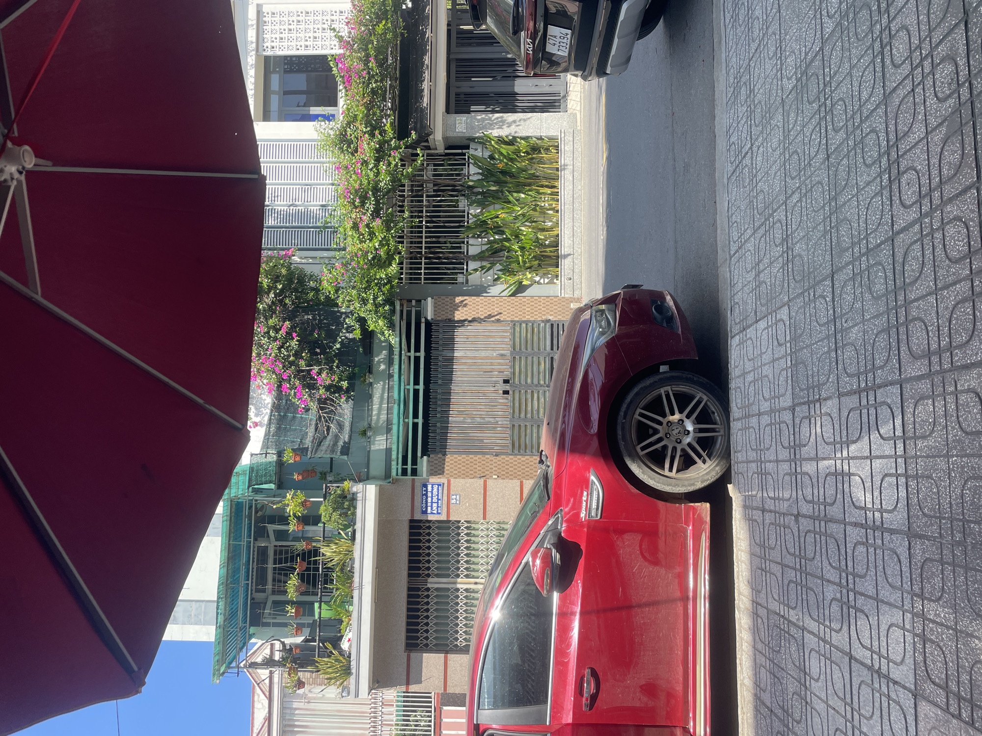This image captures a sunlit street scene in Bac Nha Trang Ward, Vietnam, viewed from beneath a large, open, deep red market umbrella, which occupies the upper portion of the frame. The foreground features a gray sidewalk composed of square pavers with an interlocking curved pattern, partially illuminated by sunlight and partially shadowed.
On the left, a significant portion of a red Chevrolet Cruze sedan is visible, parked on the street. It features multi-spoke alloy wheels and a "Sports" badge on its fender. On the right, the rear section of a black Kia SUV, bearing the Vietnamese license plate "47A 733.94", is also parked on the street. Both vehicles face away from the viewer.
Between the cars, an asphalt street separates the foreground from a row of multi-story buildings. The building immediately behind the red car has a ground floor with a light-colored facade and a white, accordion-style metal security gate. A blue rectangular sign on its wall reads "CÔNG TY BẤT ĐỘNG SẢN ANH DƯƠNG" (Anh Duong Real Estate Company) with phone numbers and the address "57A" below. The upper floors feature a balcony adorned with numerous potted plants and hanging baskets, shaded by a green awning.
Further to the right, a white metal fence covers a garden bed filled with tall, leafy green plants with some yellow blossoms. Above this fence, a dense growth of bougainvillea with vibrant pink flowers cascades over a wall. An adjacent building shows a distinct pattern of white decorative blocks and dark-framed windows on its upper floor. Another address, "62", is visible near a gray gate to the right of the garden. The background consists of a clear, cloudless blue sky, indicating bright daytime conditions. No people are visible in the scene.