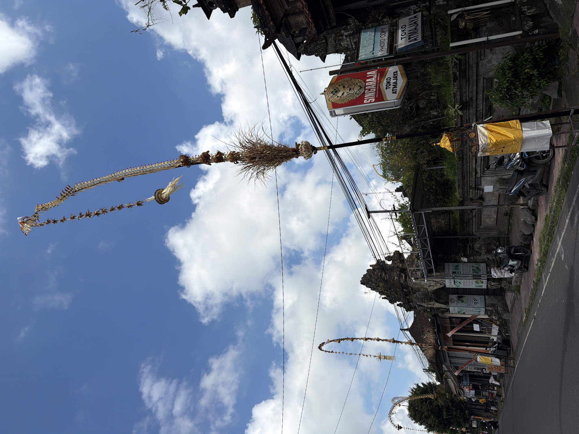 The image shows a street scene in Tampaksiring, Indonesia, with traditional Balinese decorations hanging from poles. The decorations are tall, ornate, and made of natural materials, including dried grasses and woven elements. One prominent decoration is a long, tiered structure adorned with small woven pieces and a larger bunch of dried grass at the bottom. In the background, there is a stone archway, typical of Balinese temple entrances, with carved details. Several shops line the street, with signs indicating they are "Toko" (shop). One sign reads "SINGARAJA" above "TOKO ATMAJAYA" and another is partially visible with "PHOTO COPY". The street is paved, and there are several motorcycles parked along the side. The sky is blue with scattered white clouds. The overall atmosphere suggests a cultural and possibly festive setting in a tropical environment.