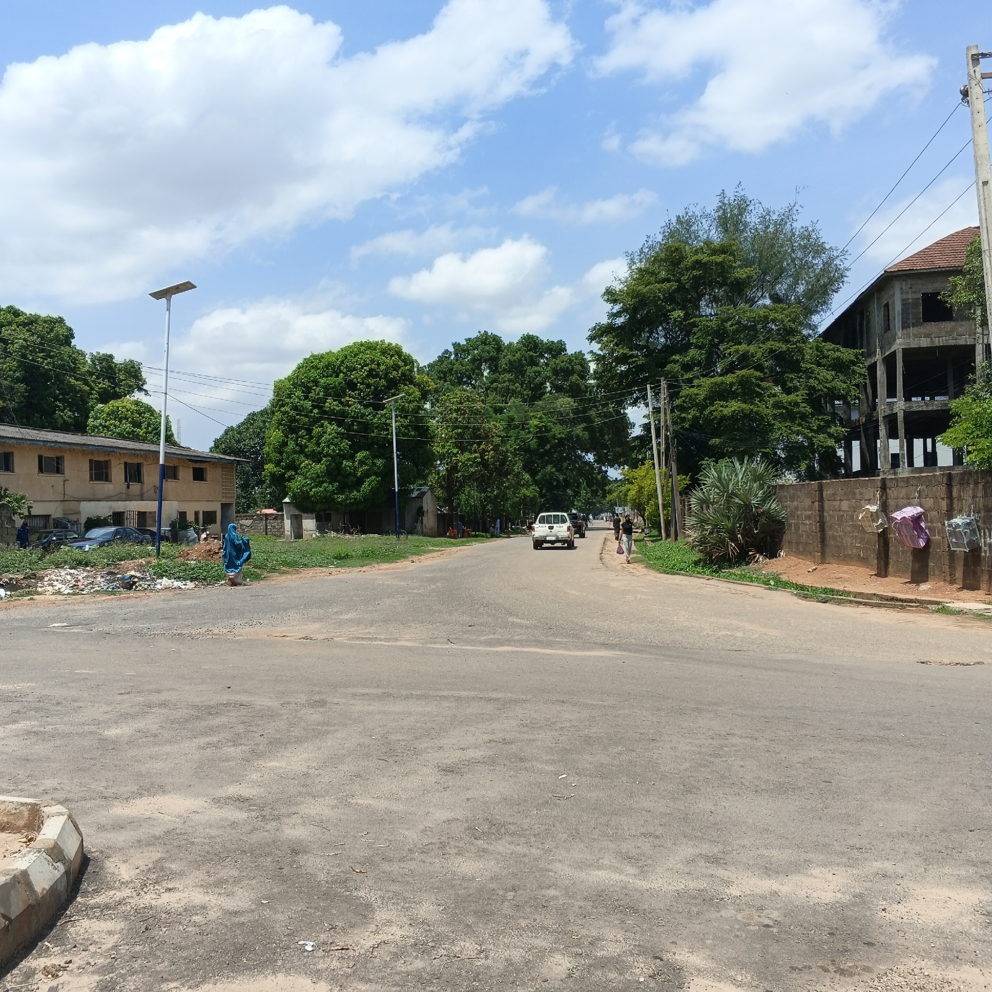 The image depicts a street scene in Shaba, Nigeria. In the foreground, a grey asphalt road occupies the majority of the frame, with a small section of a concrete curb visible on the left. The road stretches into the distance, flanked by vegetation and buildings.
On the left side of the road, a light-colored, two-story building with a dark-tiled roof is visible. Several cars are parked in front of this building, and a pile of debris is present on the ground. A person in a blue garment is standing on the side of the road, facing away from the camera. A tall, white pole with a solar panel at the top stands near this building.
Further down the road, a white pickup truck is driving away from the camera, followed by another vehicle. On the right side of the road, a brick wall runs along the property line. A large, unfinished concrete structure with multiple levels and visible beams is situated behind the wall, partially obscured by trees. Hanging on the wall are several items, including a purple and white striped fabric and two cage-like objects. A large, leafy palm-like plant grows next to the wall.
Several trees with dense green foliage are distributed throughout the scene, contributing to a lush, natural environment. The sky above is blue with scattered white clouds. Power lines and a utility pole are visible on the right side of the image. Two individuals are walking on the right side of the road in the distance.
