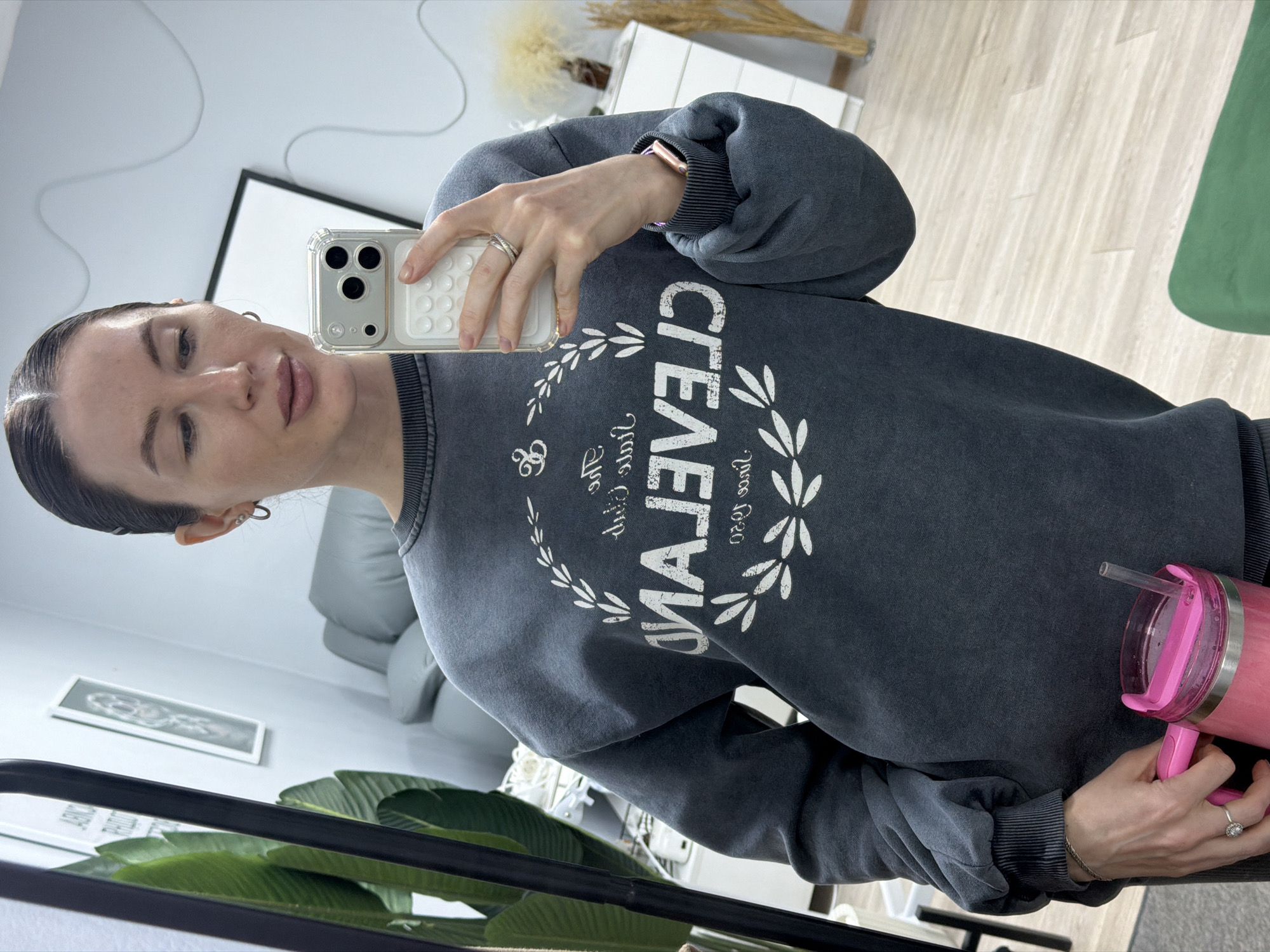 The image is a mirror selfie taken by a woman indoors in Perm, Russia. The woman, with her dark hair neatly pulled back, is wearing a dark grey, faded crewneck sweatshirt with ribbed cuffs. She holds a silver iPhone in a clear case with a white textured grip in her left hand, positioning it to capture the reflection. On her left wrist, she wears a rose gold-colored smartwatch, and two rings are visible on her left hand – a simple band and another with a prominent stone. In her right hand, she holds a vibrant pink tumbler with a silver rim, a matching pink handle, and a clear straw.
The most notable detail on her sweatshirt is the prominent white text, inverted due to the mirror, which clearly reads "CLEVELAND". This text is framed by a decorative laurel wreath design. Below "CLEVELAND", smaller inverted text is discernible, including what appears to be "since 1920".
The background reflects a modern, well-lit interior space. Features include light-colored wooden or laminate flooring and a large green potted plant in the foreground. A light grey sofa is partially visible, along with a framed piece of artwork on a white wall. Other decorative elements like dried pampas grass and a glimpse of a green furniture item contribute to the contemporary setting. The woman maintains a neutral expression, and her lips appear naturally tinted.