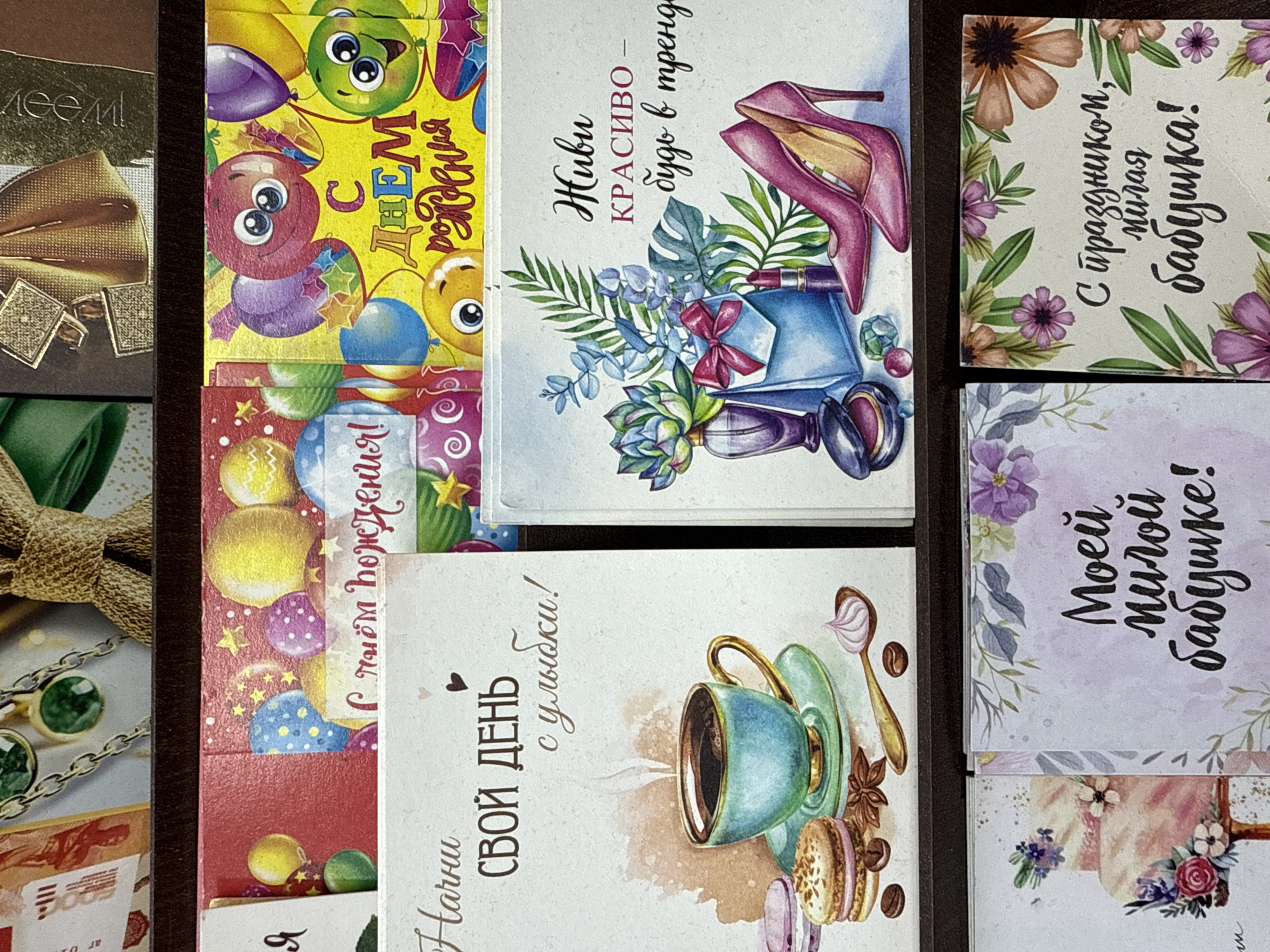 The image shows a collection of greeting cards arranged on a dark brown surface. The cards are diverse in design, with themes including birthdays, general well-wishes, and greetings for grandmothers. Some cards feature illustrated images, such as balloons, high-heeled shoes, a teacup with macarons, and floral patterns. The text on the cards is in Russian, with phrases like "Happy Birthday", "Live beautifully – be in trend", "Start your day with a smile!", "To my dear grandmother!", and "Happy holiday, dear grandmother!". The cards appear to be stacked, with some overlapping, indicating they may be part of a collection or display. It is not possible to confirm the specific location of Nizhnevartovsk, Russia from the contents of the image.