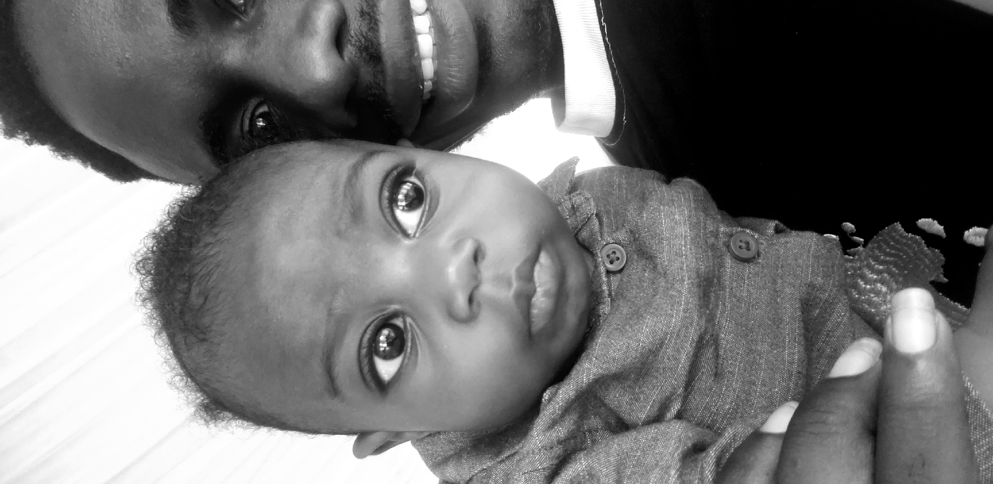 A black and white, close-up, head-and-shoulders portrait shows a dark-skinned baby being held by a dark-skinned adult male. The adult is on the right side of the frame, and the baby is on the left, nestled against the adult's chest. The baby's face is the primary focus, with large, dark eyes looking off to the right. The baby is wearing a collared, button-up shirt, the top button visible. The adult's face is partially visible, with a mustache and a wide smile showing teeth. The adult is wearing a dark shirt with a white collar visible. The background is bright and indistinct, suggesting an outdoor setting with a light-colored structure or fabric. The image appears to be a personal photograph taken in Narayi, Nigeria.