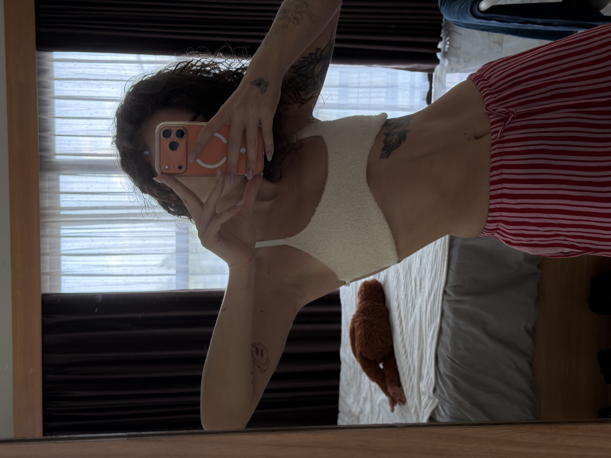 A young woman with curly brown hair is taking a mirror selfie in a bedroom. She is wearing a white fuzzy bralette and red and pink striped pajama pants. Her phone, which is in an orange case, is held up to her face, obscuring it. She has tattoos on her arms and torso. In the background, a bed is visible with a brown stuffed animal lying on it. Dark brown curtains are drawn on the left side of the window, and sheer white curtains are on the right. The room appears to be in a hotel or rental property in North Kuta, Indonesia.
