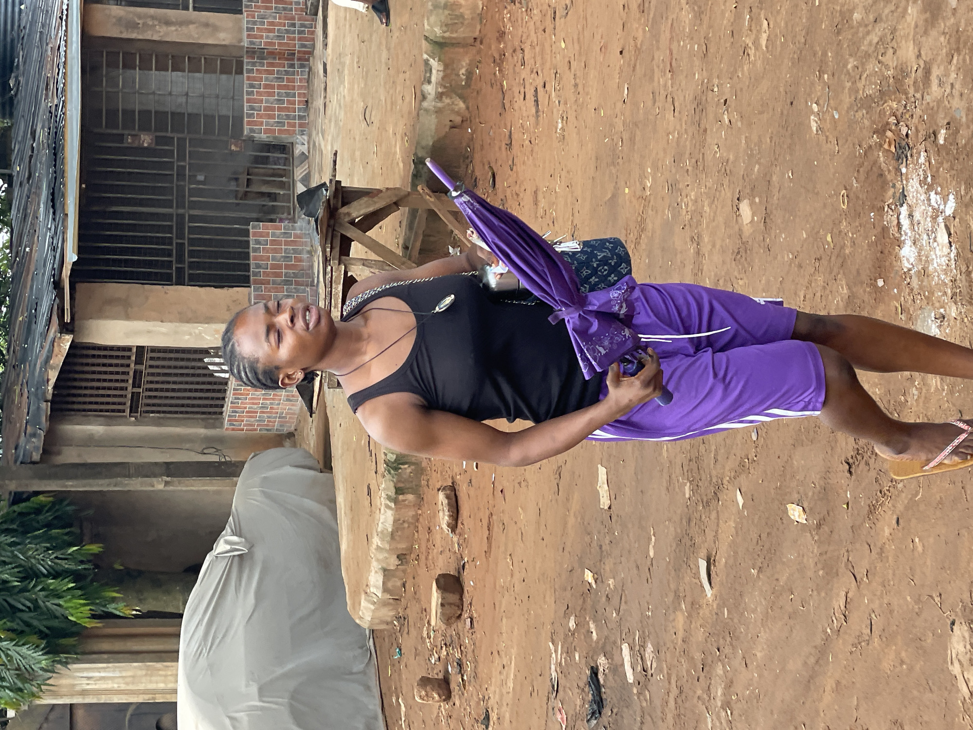 A woman stands outdoors in what appears to be Onitsha, Nigeria, based on the environmental conditions and architecture visible. She is wearing a black tank top and purple shorts with white stripes down the sides. She holds a closed, purple umbrella and has a small handbag slung over her shoulder. The background includes a building with brickwork and metal barred windows, as well as a car covered by a tarpaulin. The ground is dirt with scattered debris.
