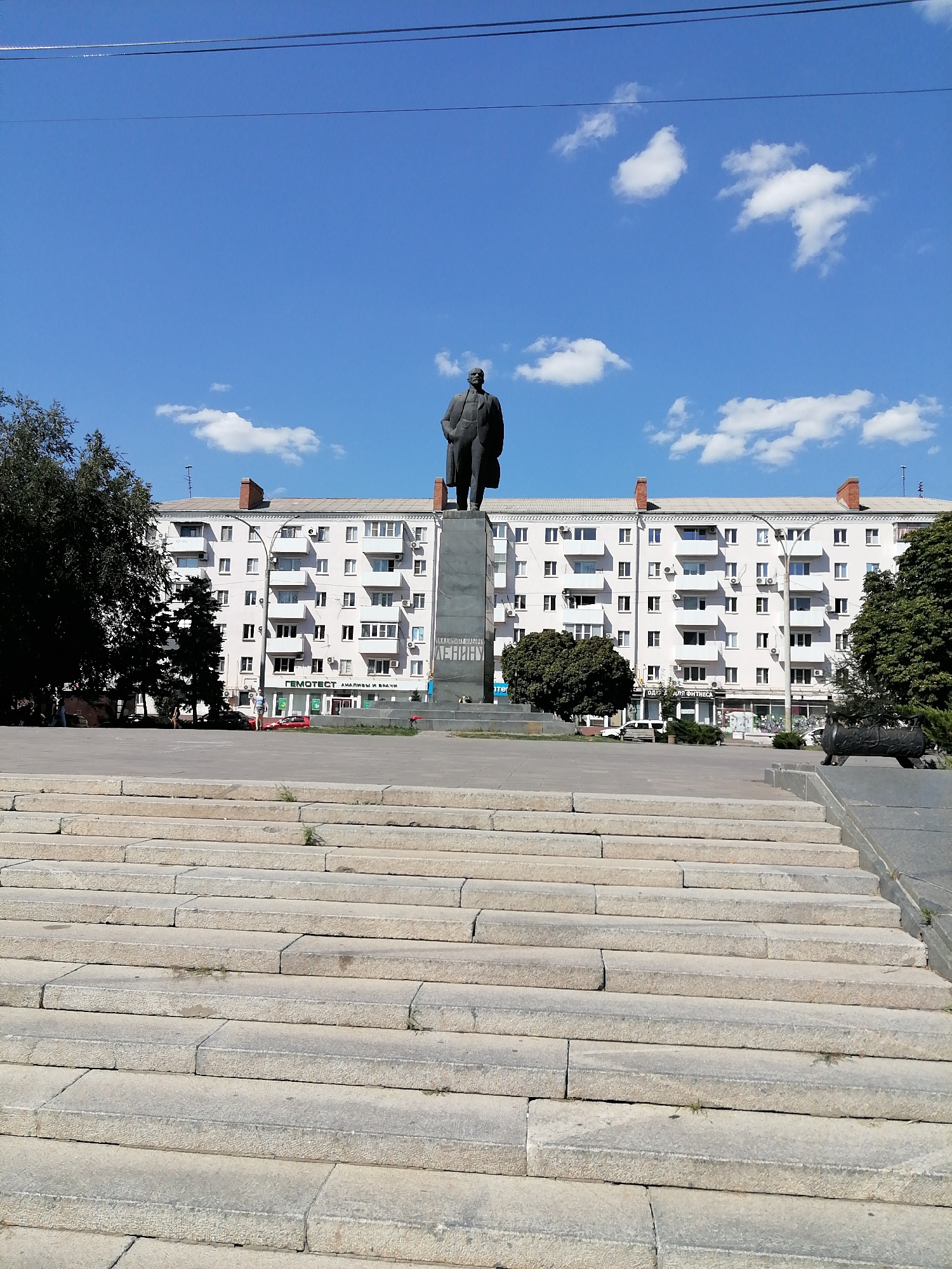 The image shows a view of a city square with a statue of Vladimir Lenin in Rostov-on-Don, Russia. In the foreground, there are large stone steps. Behind the steps, the Lenin statue stands tall on a pedestal. The statue is made of dark material and depicts Lenin in a suit, with his right hand in his pocket and his left hand extended slightly forward. The pedestal has inscriptions on it, one of which appears to read "LENIN."
In the background, a large, light-colored apartment building with multiple stories and many windows stretches across the frame. The building has a flat roof with chimneys. Trees are visible on the left and right sides of the square, providing some shade. The sky is blue with scattered white clouds. A few cars are parked on the street in front of the apartment building. A woman is walking on the sidewalk in front of the building. To the right of the statue, there is a dark-colored sculpture resembling a cannon. The overall scene suggests a public space in a city, likely a monument or historical site.