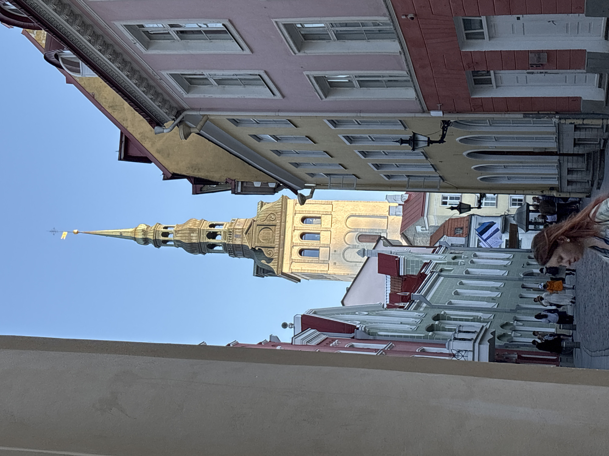 The image depicts a narrow cobblestone street in Tallinn Old Town, Estonia, framed by historic buildings under a clear blue sky. In the background, the distinctive, light-colored spire and tower of St. Olaf's Church (Oleviste kirik), topped with a golden finial and a small golden pennant, rises prominently.
On the left side of the street, a sequence of multi-story buildings features pitched red roofs and facades in shades of red, light green, and cream. Architectural details include white window frames, some small balconies, and decorative cornices. An Estonian flag (blue, black, and white horizontal stripes) is visible hanging from one of these buildings further down the street. The immediate foreground on the left is obscured by a plain, light beige wall, suggesting a building corner or archway.
On the right side of the street, buildings exhibit various colors and architectural styles. The closest building has a pink upper facade with white windows and a reddish lower section with two white paneled doors. Adjacent to it, a multi-story yellow building features a row of arched windows on its lower level and rectangular windows above, adorned with ornate black wrought-iron street lamps. Further back, another yellow building with a red roof is partially visible.
The street itself is paved with dark cobblestones. Several individuals are present in the middle ground, walking along the street. One woman with reddish-brown hair is partially visible in the bottom right foreground, facing away from the camera. The scene is illuminated by natural light, highlighting the church tower and casting some shadows on the street.