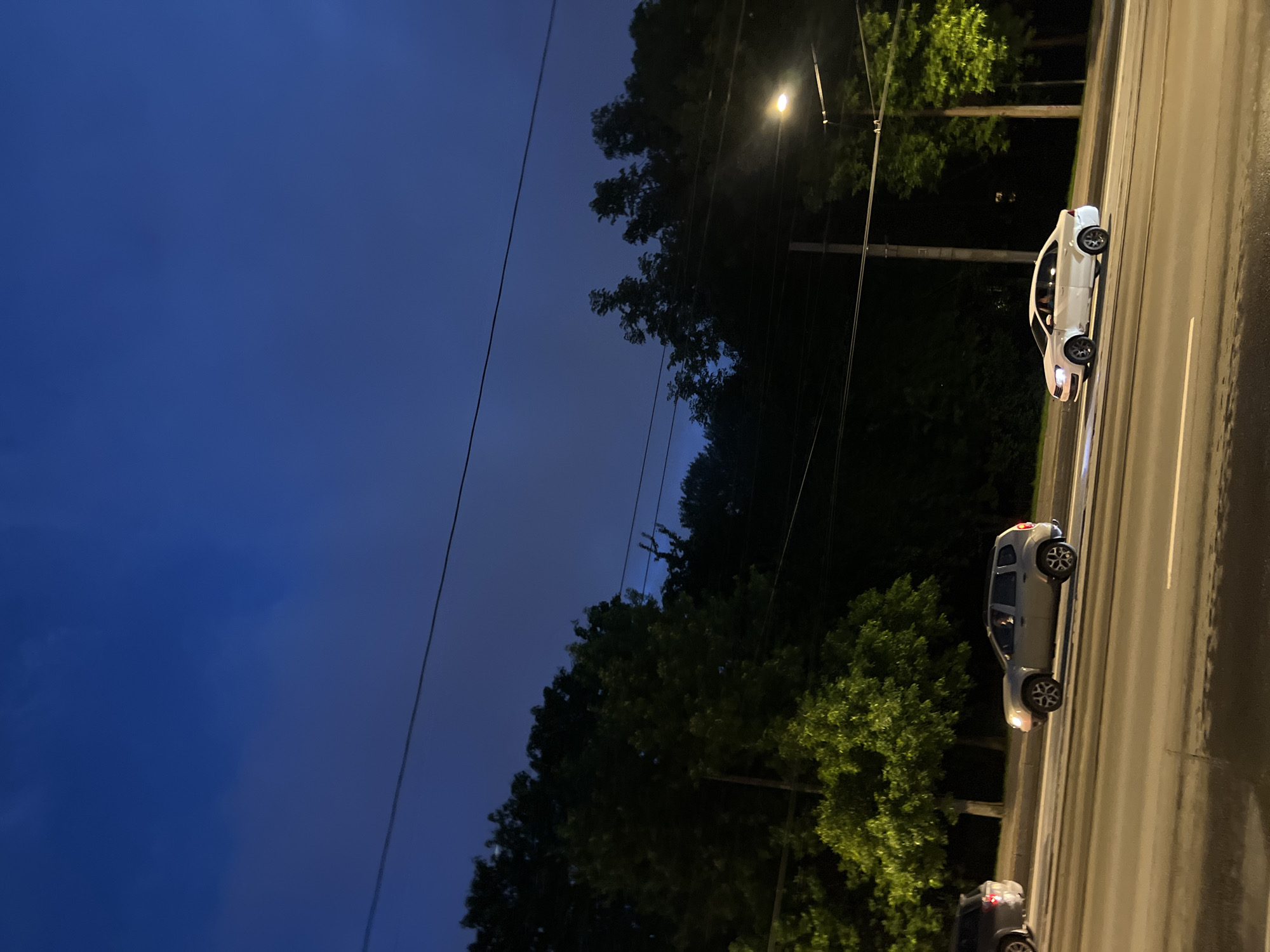 The image shows a street scene in St. Petersburg, Russia, at dusk. The sky is a deep blue, transitioning to darker shades as night falls. Several cars are driving on a multi-lane road. In the foreground, a light-colored sedan is in the right lane, and a light-colored SUV is in the left lane. Further back, another dark-colored SUV is partially visible in the left lane. Trees line the side of the road, and a street light is illuminated on the right side of the frame. Electrical wires are visible across the top of the image, suggesting the presence of tram lines or street lighting infrastructure. The road surface appears wet, indicating recent rain or damp conditions.