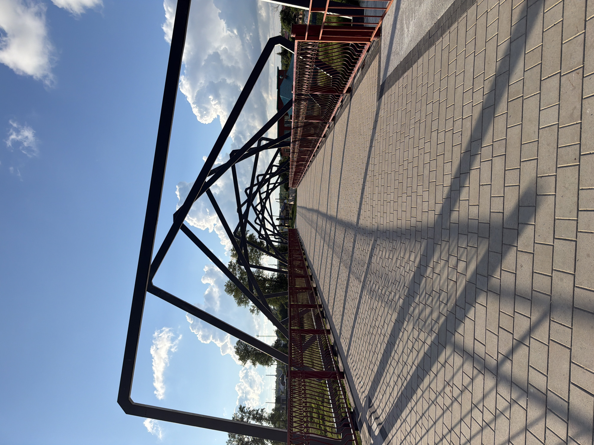 This image shows a pedestrian bridge in Solikamsk, Russia. The bridge has a modern design with a dark metal frame and a red metal railing. The walkway is paved with light-colored rectangular bricks. Shadows from the bridge's structure are cast across the walkway, indicating it is daytime. The sky is blue with large, fluffy white clouds, suggesting fair weather. Trees and some buildings are visible beyond the bridge, hinting at an urban or suburban setting near a body of water, possibly a river or canal, judging by the presence of a railing on the far side of the bridge. The scene appears peaceful and empty, with no people or animals visible. The image is taken from a low angle, looking along the length of the bridge towards the horizon. The sun is likely low in the sky, casting long shadows.