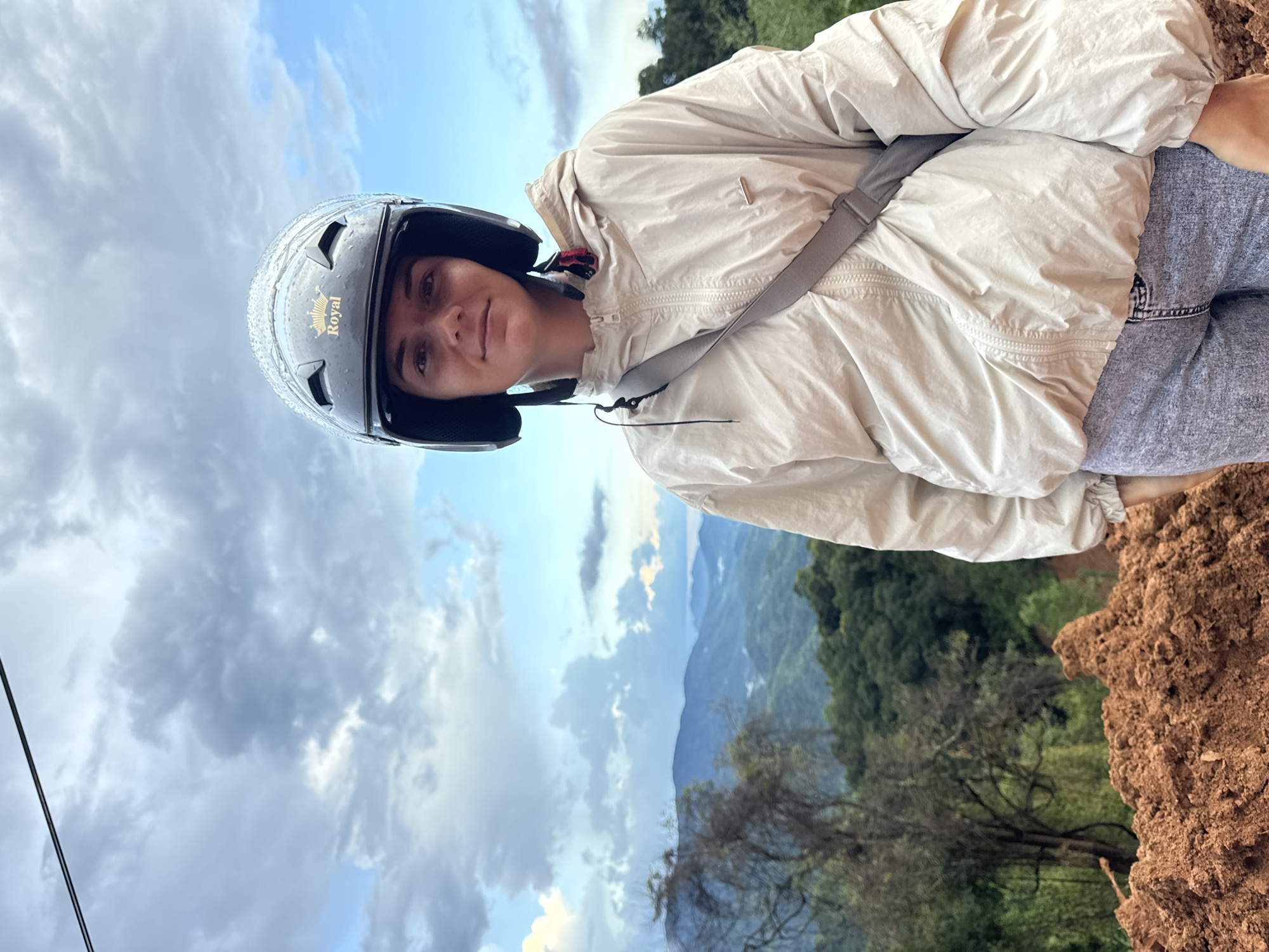 A young woman, wearing a black motorcycle helmet with a "Royal" logo and a light beige jacket, stands in front of a scenic mountain landscape. She is looking directly at the camera, with a slight smile. The background consists of lush green hills and mountains, under a cloudy sky with patches of blue. In the foreground, there is a mound of reddish-brown soil. A thin black wire is visible at the top of the frame. The location is identified as Nao Sẻ, Vietnam.