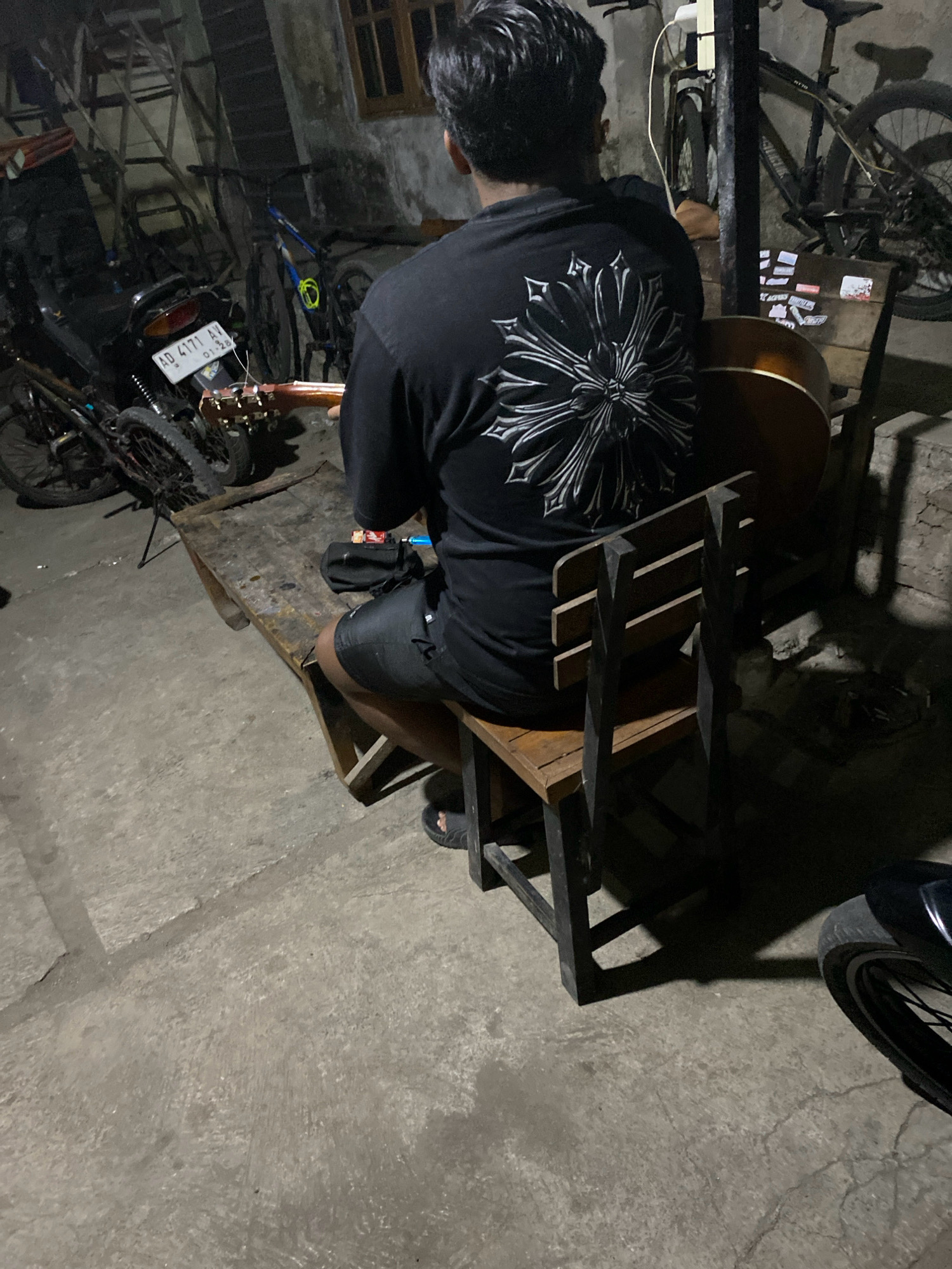 A man sits on a wooden chair and plays an acoustic guitar in what appears to be a garage or outdoor workshop in Pondok, Indonesia. He is wearing a black t-shirt with a large silver floral design on the back and black shorts. The guitar's body is visible resting on a wooden table. Several motorcycles and bicycles are parked in the background, some leaning against walls. The ground is a concrete surface with visible cracks and stains. A wooden table with a black pouch on it is positioned in front of the man. The scene is dimly lit, suggesting it is either nighttime or the area is enclosed and lacks sufficient lighting.