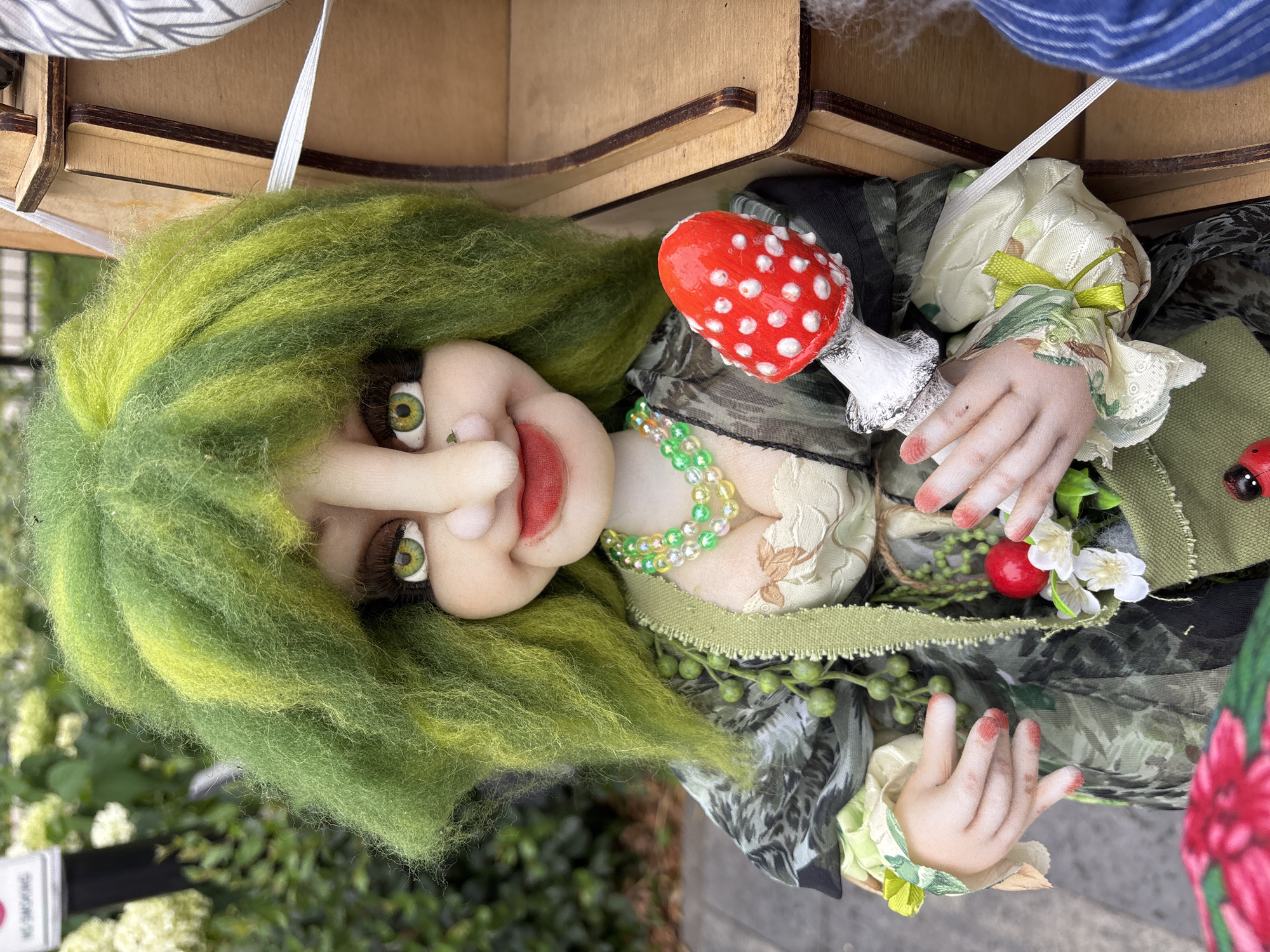 A doll, likely handcrafted, is depicted outdoors. The doll has bright green, spiky hair and large, expressive eyes with prominent eyelashes and eyebrows. Its face is characterized by rosy cheeks, a wide red smile, and a slightly upturned nose. It wears a green and black patterned dress with floral accents, a beaded necklace, and holds a red mushroom with white spots. The doll is positioned in front of a wooden display structure, and in the background, there is lush green foliage and a white sign. The overall impression is of a whimsical, possibly fantasy-themed figure. The image was taken in Zvenigorod, Russia.