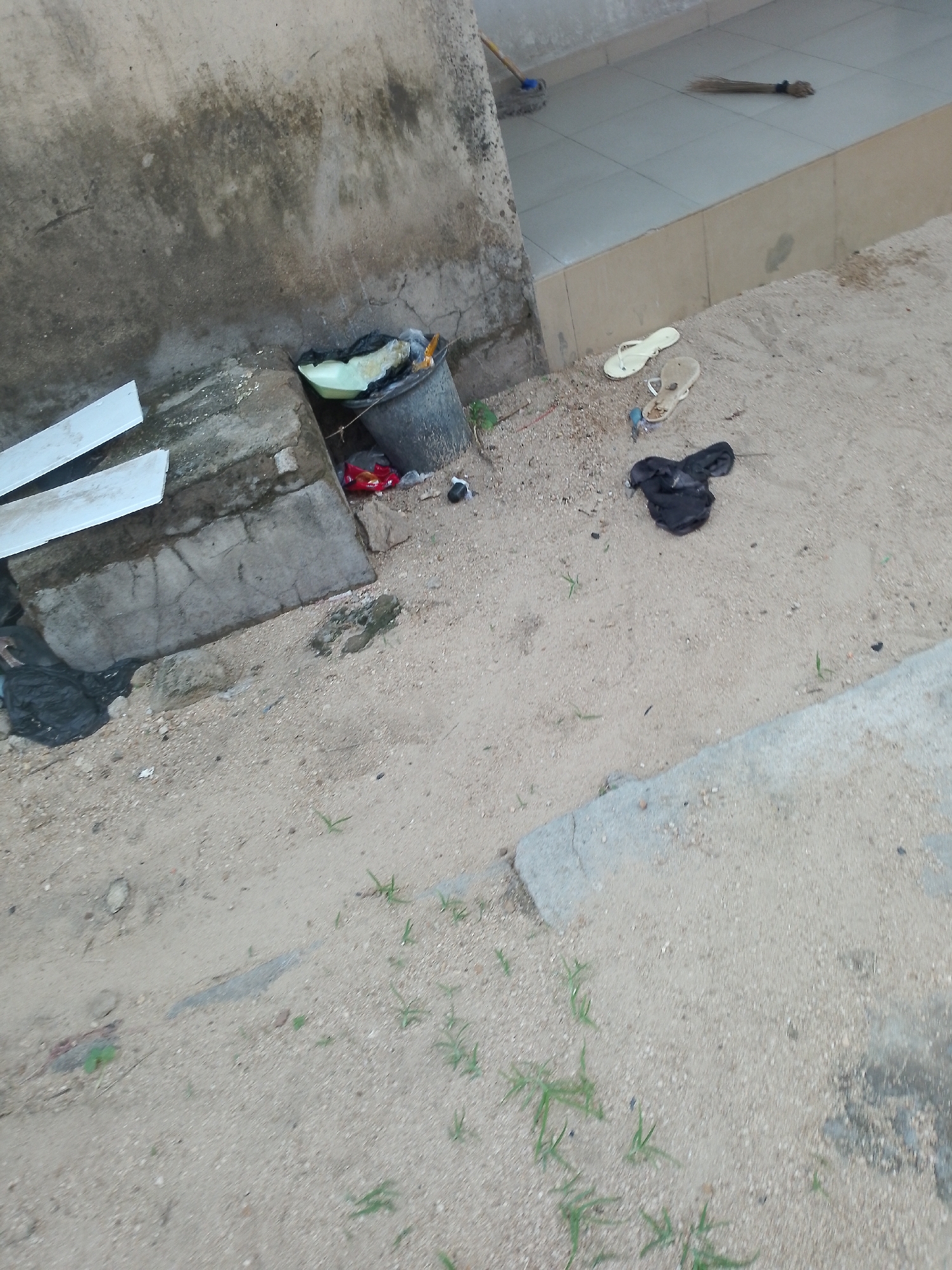 In Jauro Yinu, Nigeria, a weathered, textured concrete wall forms the backdrop of a scene of disarray. Below the wall, a black plastic bin lies on its side, spilling its contents of a light green plastic basin and some crumpled red packaging. Next to the bin, a black plastic bag is partially visible. In the sandy ground, a pair of light beige flip-flops lies next to a single black, cloth item. To the left, two long, flat white objects are propped against a concrete structure. Further to the right, on a tiled surface, a broom lies horizontally. The overall environment is dusty and appears unkempt.