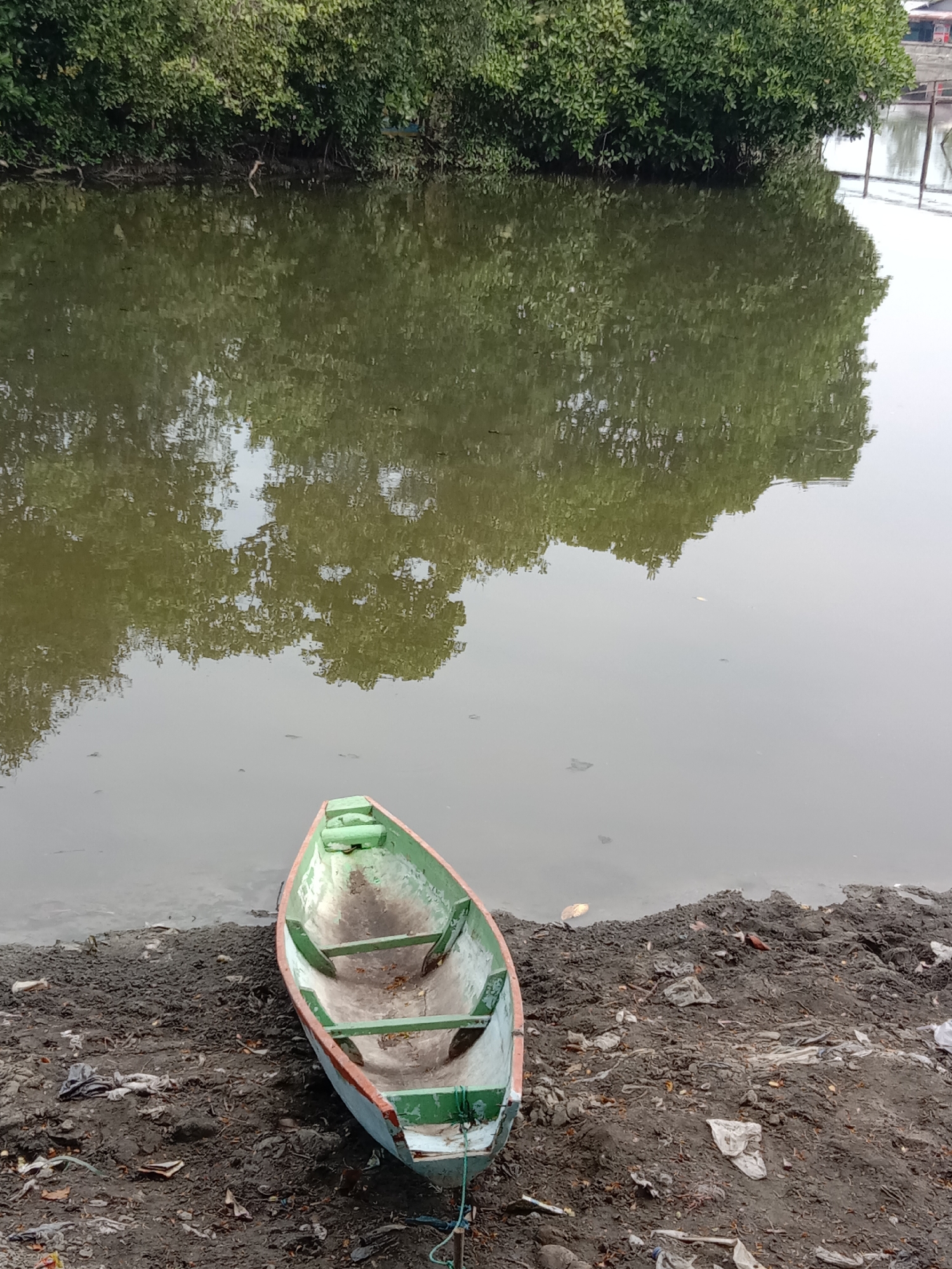 An unpainted wooden boat, lined with green and orange paint, sits on a muddy bank littered with trash along a body of water. The water reflects trees and sky. The scene is located in Lhokseumawe, Indonesia.