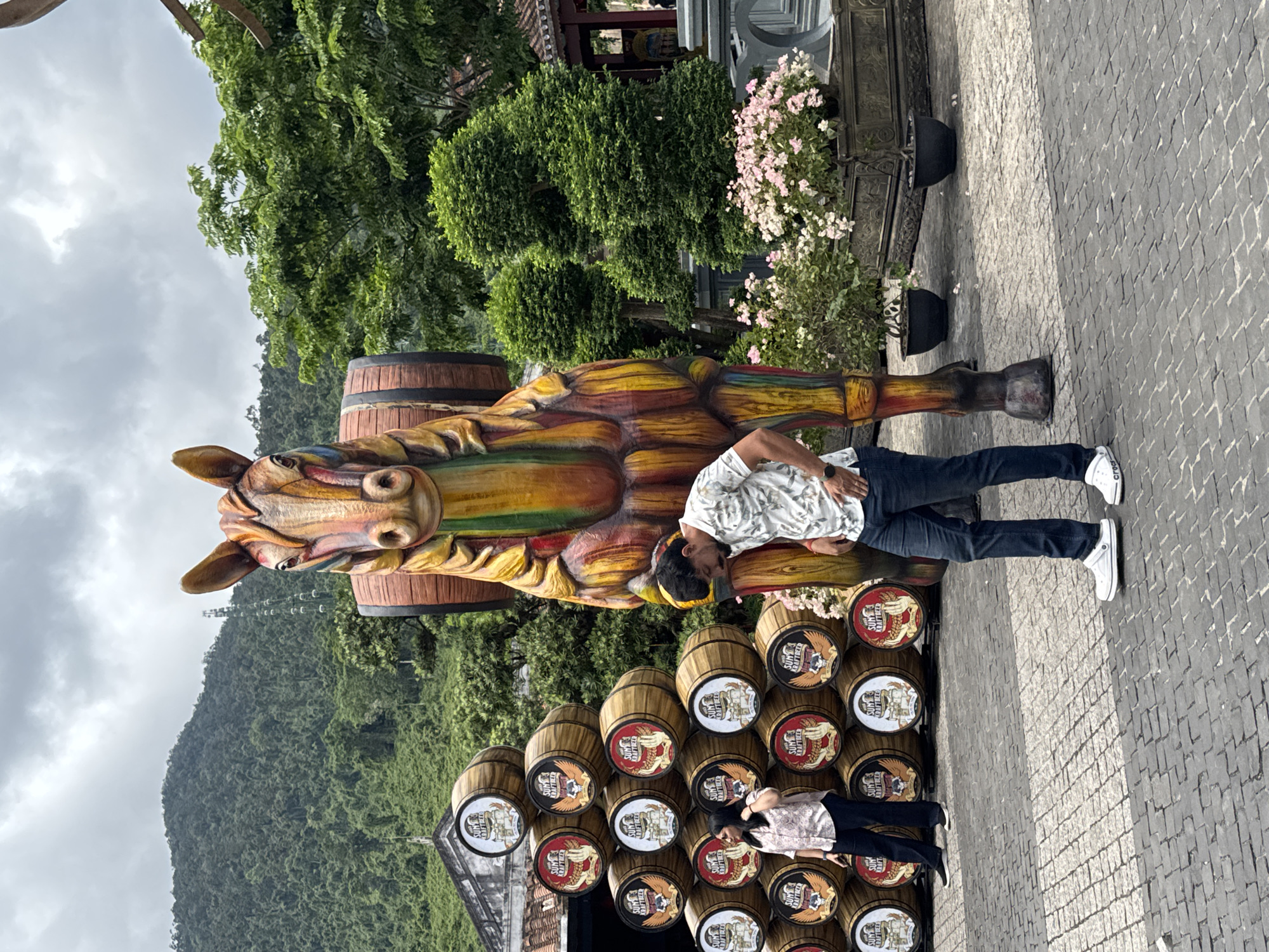 The image captures two people posing near a large, colorful wooden horse statue erected on a foundation of barrels, likely in a public area. The statue is the focal point, vividly painted with yellows, reds, and greens. The barrels beneath the statue feature branding with the words "SUN KRAFTBEER". One person, dressed in a light patterned shirt and dark jeans, is leaning against the body of the horse, while another person is positioned near the base of the barrels, wearing a light-colored shirt and dark pants.
In the background, a lush mountain range, topped with trees, stretches across the horizon under an overcast sky. To the side of the statue, manicured shrubs and flower planters line a stone-paved area. The architecture in the backdrop appears to be from a theme park. Considering the theme and architecture, this location is likely within Sun World Ba Na Hills in Đà Nẵng, Vietnam.