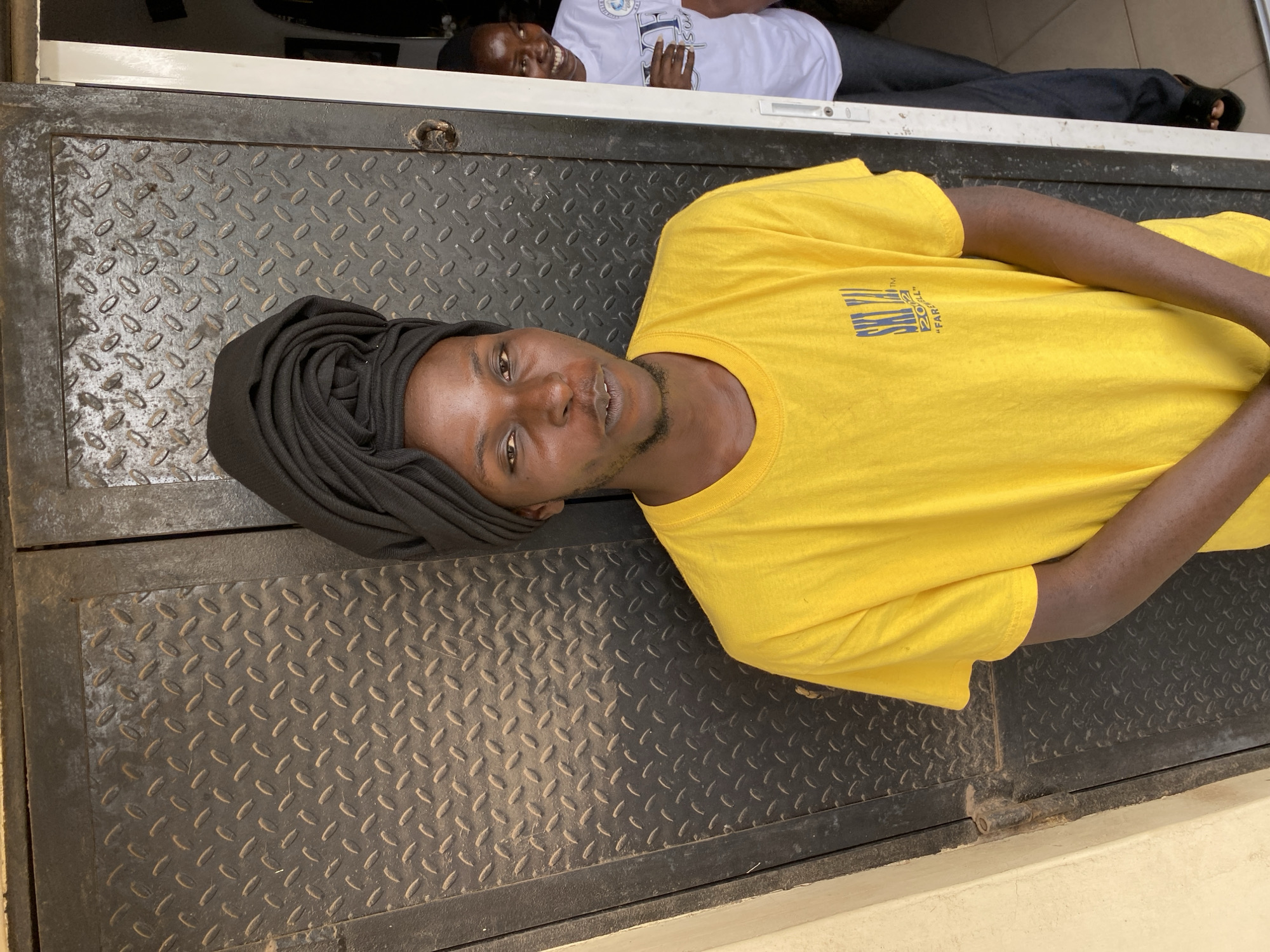 Two men are lying on a diamond-plated metal surface. One man is wearing a yellow t-shirt and a dark-colored head covering. The second man is wearing a white t-shirt. The location is Jos, Nigeria.