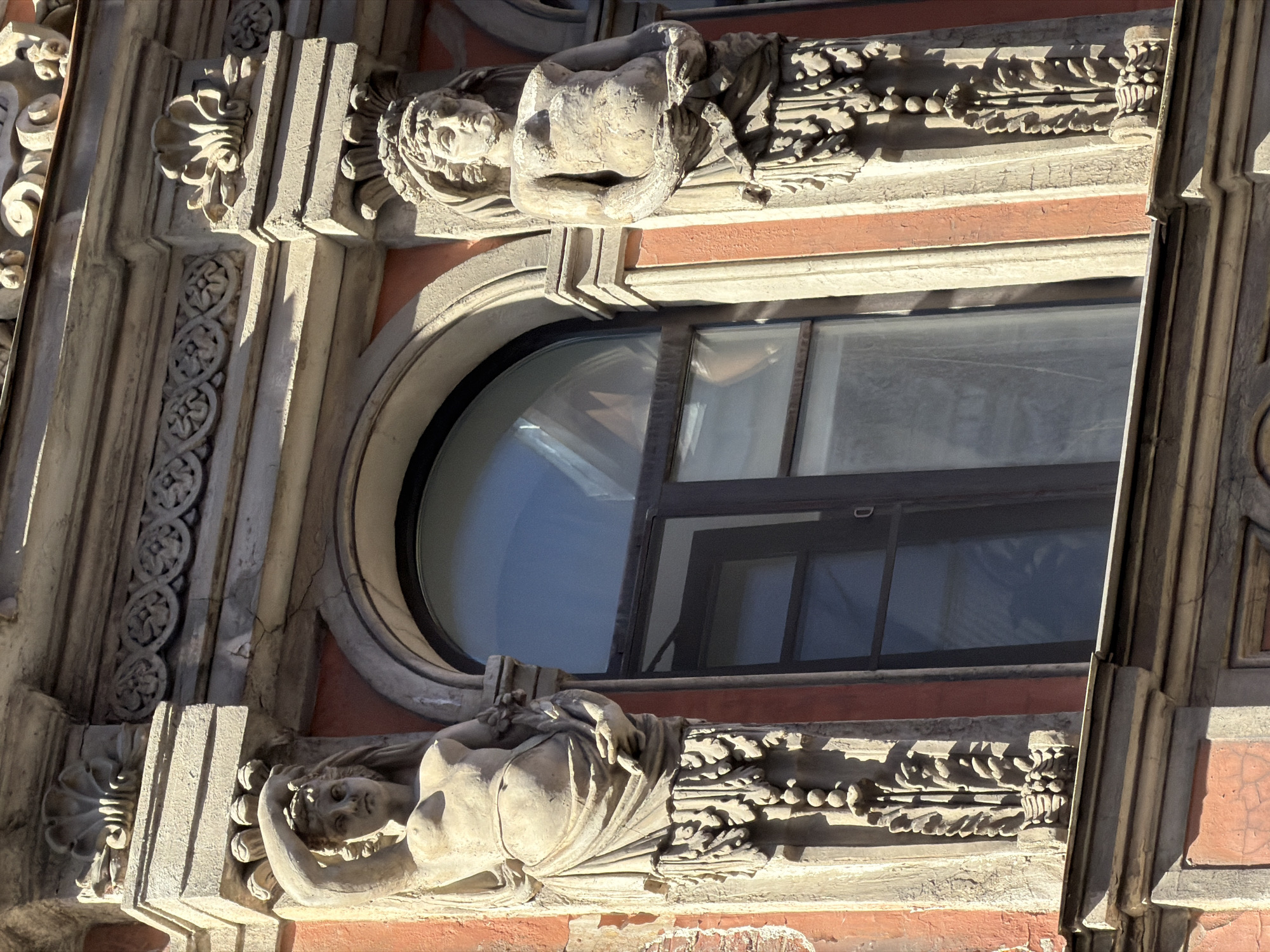 This is a close-up, slightly angled shot of architectural details on the facade of a building in Saint Petersburg, Russia. The image showcases a large, oval-shaped window with a dark frame, reflecting a bright sky. Above and below the window are ornate stone carvings.
The carvings depict stylized human figures, likely female, incorporated into decorative friezes. The figure above the window appears to be reclining, with her upper body exposed and her head turned to the side. She is adorned with drapery and is positioned above a band of intricate carvings that resemble foliage or floral motifs.
Similarly, the figure below the window is also lying down, partially covered by drapery, and her head is resting on her arm, looking downwards. This figure is also situated above a decorative frieze. Both figures are made of stone and show signs of age and wear, with some chipping and erosion visible. The building's facade is characterized by these rich, sculpted elements, suggesting historical or classical architectural influences.
The lighting indicates it is daytime, with strong sunlight casting distinct shadows on the stone carvings, highlighting their three-dimensional forms. The sky reflected in the window suggests clear weather. No visible text is present in the image. The overall impression is one of artistic craftsmanship and the weathered beauty of historic architecture.