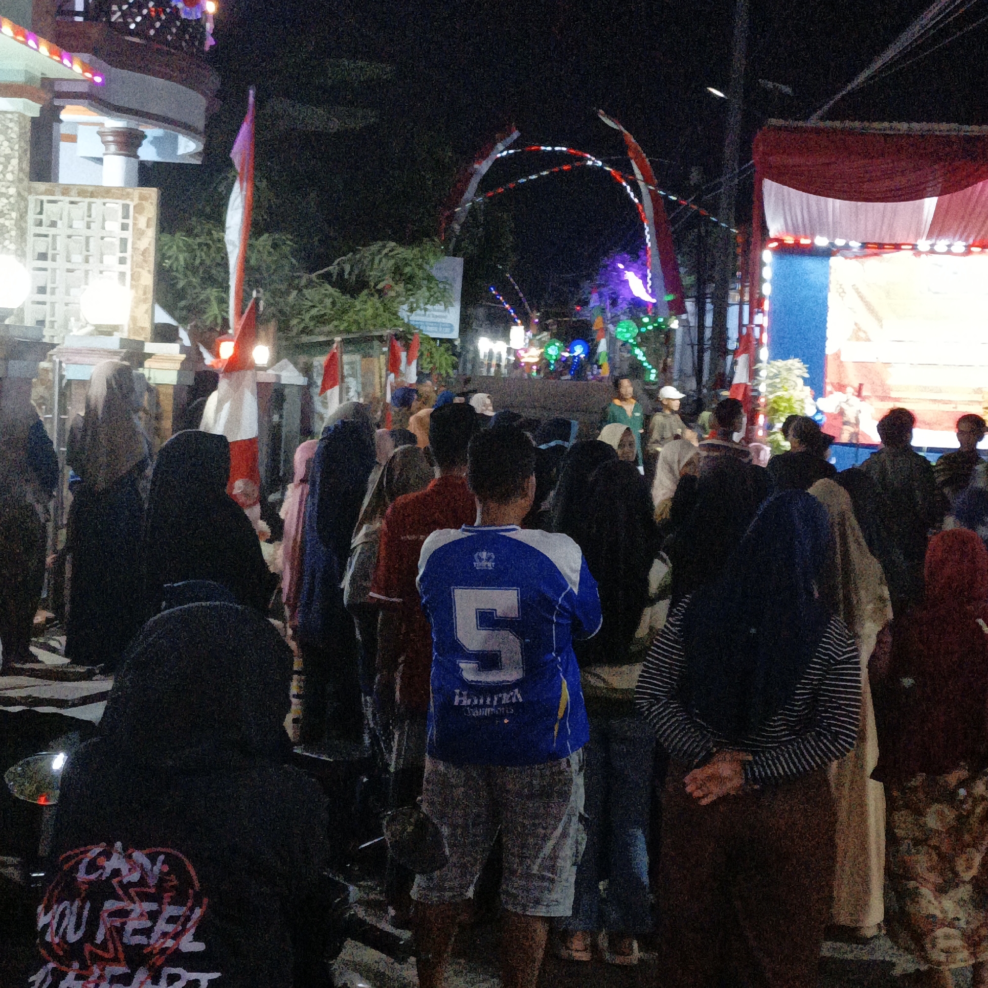 This image captures a nighttime scene in Tanahbaya, Indonesia, likely during a local celebration or event. A crowd of people, predominantly wearing hijabs or head coverings, are gathered in a street illuminated by festive decorations. Red and white flags, possibly Indonesian flags, are prominently displayed, along with colorful string lights that arc across the street. In the background, a stage or platform is visible, draped with red and white fabric, suggesting a performance or presentation is taking place. The overall atmosphere appears to be one of community gathering and festivity.