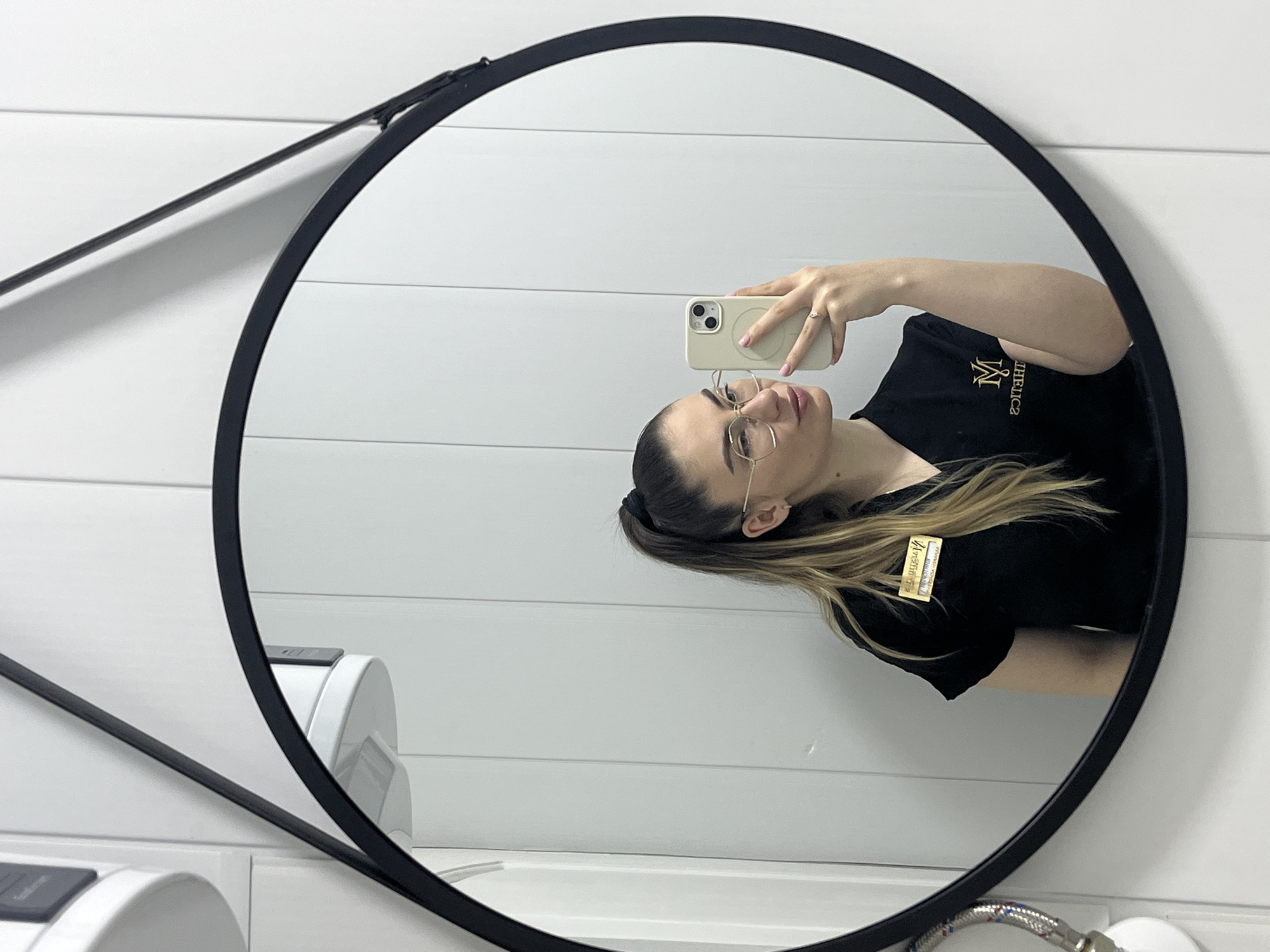 A mirror reflects a woman holding a phone in a restroom. The woman is wearing a black t-shirt and glasses. A name tag is visible on her shirt. The restroom features white walls with horizontal paneling, a black round mirror with a support arm, hand dryers, and a faucet. The location is Unknown, Unknown.