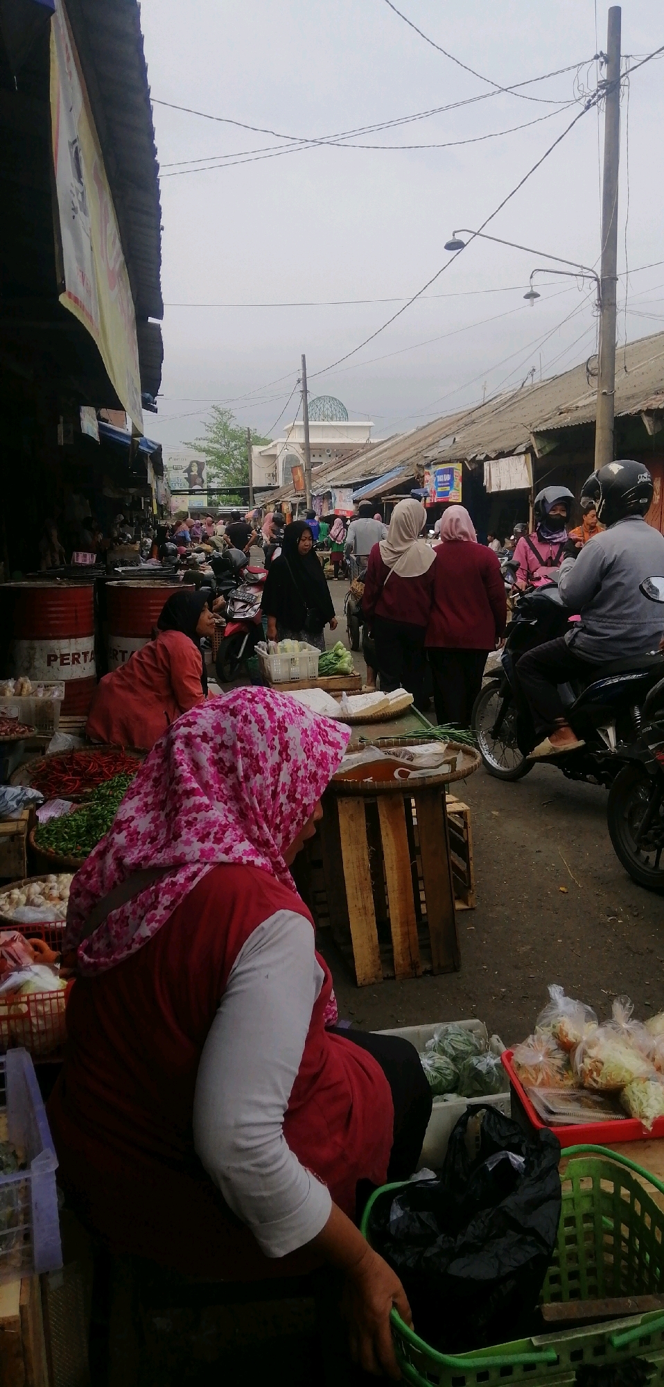 The image depicts a street market scene in Ajibarang, Indonesia, likely on a cloudy day. In the foreground, a person wearing a red shirt and a pink floral hijab is seated, facing away from the camera, with their hands near a green basket containing a black plastic bag. In front of them, there are various vegetables and produce displayed on wooden crates and in baskets, including chili peppers and what appear to be onions or garlic. To the left, two large red barrels with the word "PERTA" are visible.
Further into the market, several other individuals are present, mostly women wearing hijabs, engaged in shopping or selling. Some are standing, others are walking. A mosque with a dome is visible in the background, suggesting the location is within a community. Several motorcycles are parked and one is being ridden by a person in a helmet. The market stalls are constructed from various materials, including wood and corrugated metal, and there are overhead wires and streetlights. The ground is paved, appearing to be asphalt. The overall atmosphere suggests a bustling local marketplace.