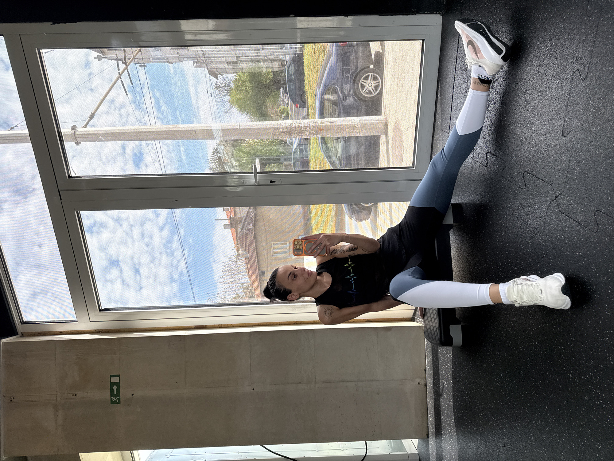 This image, taken in Pleven, Bulgaria, captures a woman engaged in an exercise or stretching routine inside what appears to be a gym or fitness studio. She is lying on her back on a black, textured interlocking floor, with her left leg extended upwards, braced against a black step bench. Her right leg is bent. She is dressed in athletic wear, including two-toned leggings (grey/blue and white), a black tank top featuring a colorful heart-rate line graphic, and white athletic shoes, one of which displays a Nike logo and the word "RUN." White socks with partially visible, reversed text "SPORT" and "ATHLETIC" are also worn.
The woman is actively taking a selfie with an orange-cased smartphone, which shows an Apple logo, held in her right hand above her face. Her expression is focused, with a slight upward gaze towards the phone, suggesting she is monitoring her pose or framing the shot.
Large windows behind her reveal a bright daytime outdoor scene. The sky is blue with scattered white clouds, indicating clear weather. Through the window, a street with parked vehicles, including a dark SUV and a blue sedan, green grass, distant buildings, and utility poles with wires are visible. The windows are equipped with a fine mesh screen. Inside, below the window, a concrete wall features a green emergency exit sign with an upward-pointing arrow and a stylized human figure.