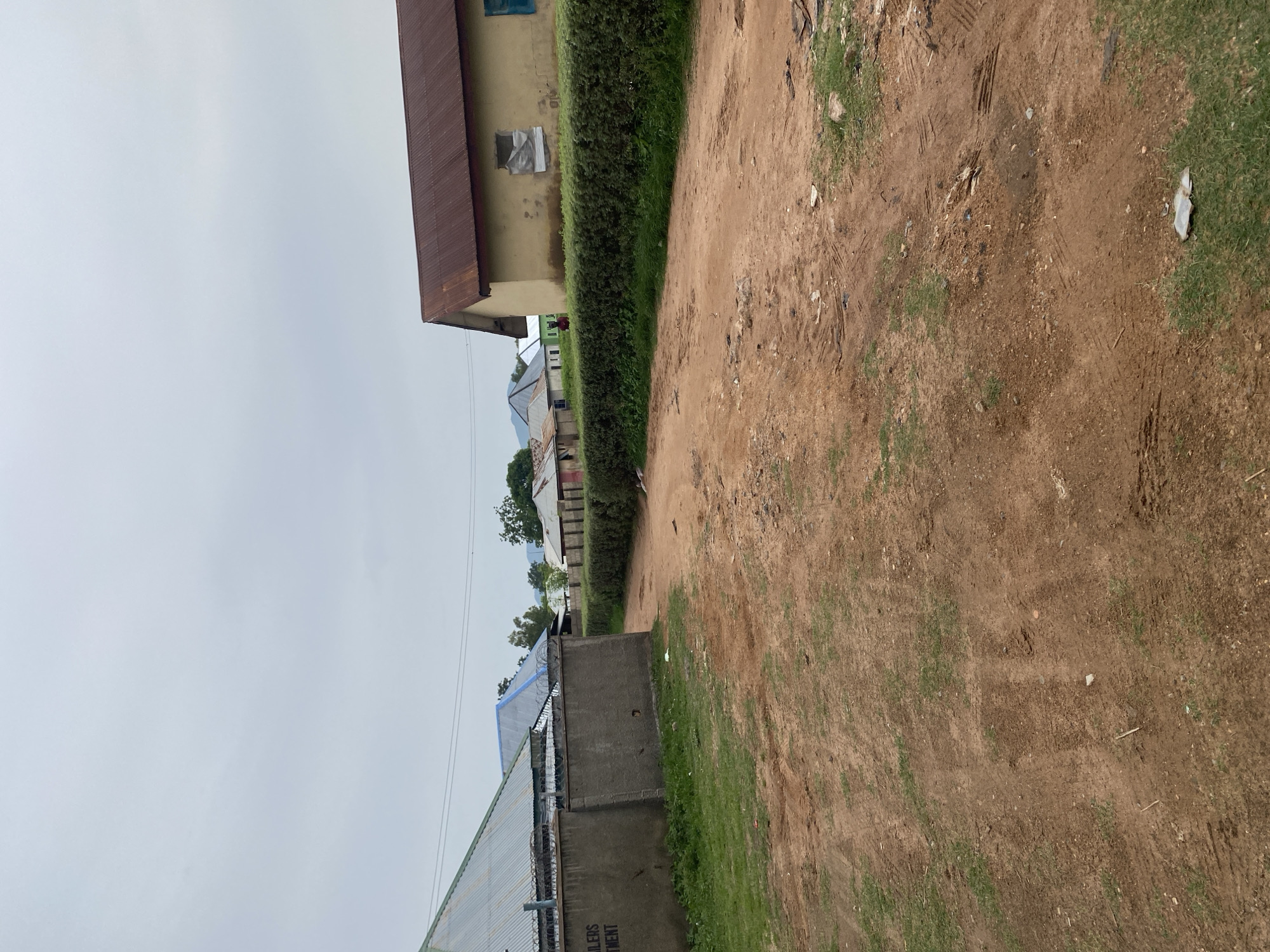 The image depicts a dirt road in Jauro Yinu, Nigeria, with several buildings lining the right side of the road. The buildings appear to be single-story structures with corrugated metal roofs, some painted in shades of yellow and brown, while others have tin roofs. A dense hedge runs along the front of the properties, obscuring the lower portions of the buildings. On the left side of the road, a concrete wall topped with barbed wire is visible, with a building behind it that also has a corrugated metal roof. The road surface is predominantly red earth, marked with tire tracks and some scattered debris. The sky is overcast with a light gray hue. A single individual, appearing as a silhouette from this distance, is standing near one of the buildings on the right. The environment suggests a rural or semi-urban setting.