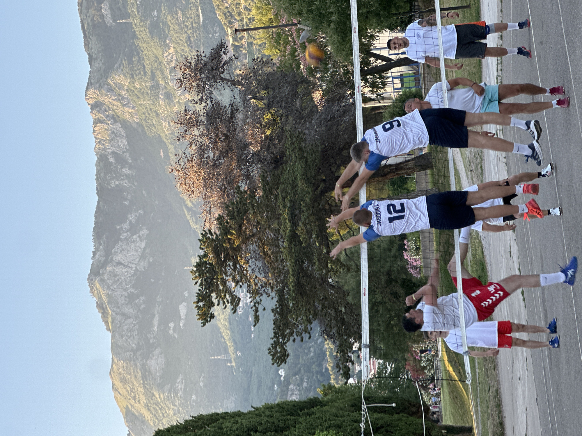 A group of men are playing a game of beach volleyball outdoors in Budva, Montenegro. The court is paved with asphalt and is bordered by a volleyball net. In the background, a range of lush green mountains rises under a clear blue sky. The players are dressed in athletic attire, with some wearing jerseys and shorts, while others are in t-shirts and shorts. One player in red shorts is captured mid-air, performing a powerful kick. Another player in a white jersey with the number 12 on it is also jumping, with his arms raised as if to hit the ball. The game appears to be in full swing, with one player in a white jersey with the number 9 on it reaching to block a shot, while another player in a white t-shirt and light blue shorts watches intently. In the distance, trees and some buildings are visible, blending into the mountainous landscape. The scene is illuminated by the warm glow of the sun, casting soft shadows on the court.