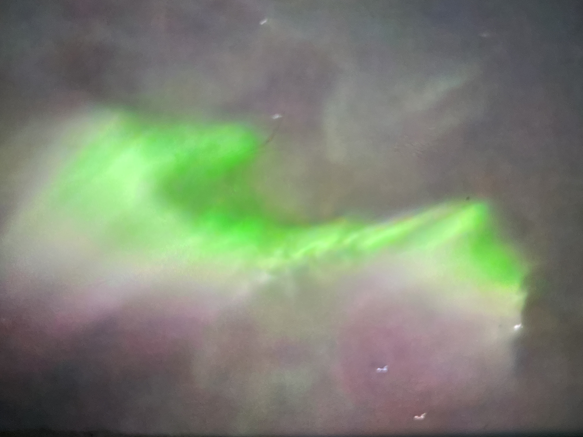 The image captures a vibrant green aurora borealis phenomenon against a dusky, purplish sky. The aurora is characterized by luminous streaks and swirls, with a prominent, bright green band dominating the central portion of the frame and extending downwards. Subtle hints of pink and lighter green are visible in the background, suggesting other atmospheric effects or clouds. A few small, faint white specks, possibly distant lights or stars, are scattered throughout the scene. The overall impression is one of a dynamic and awe-inspiring natural light display in a dark, open environment. The location is specified as Unknown, Unknown, indicating the setting is not precisely identified.