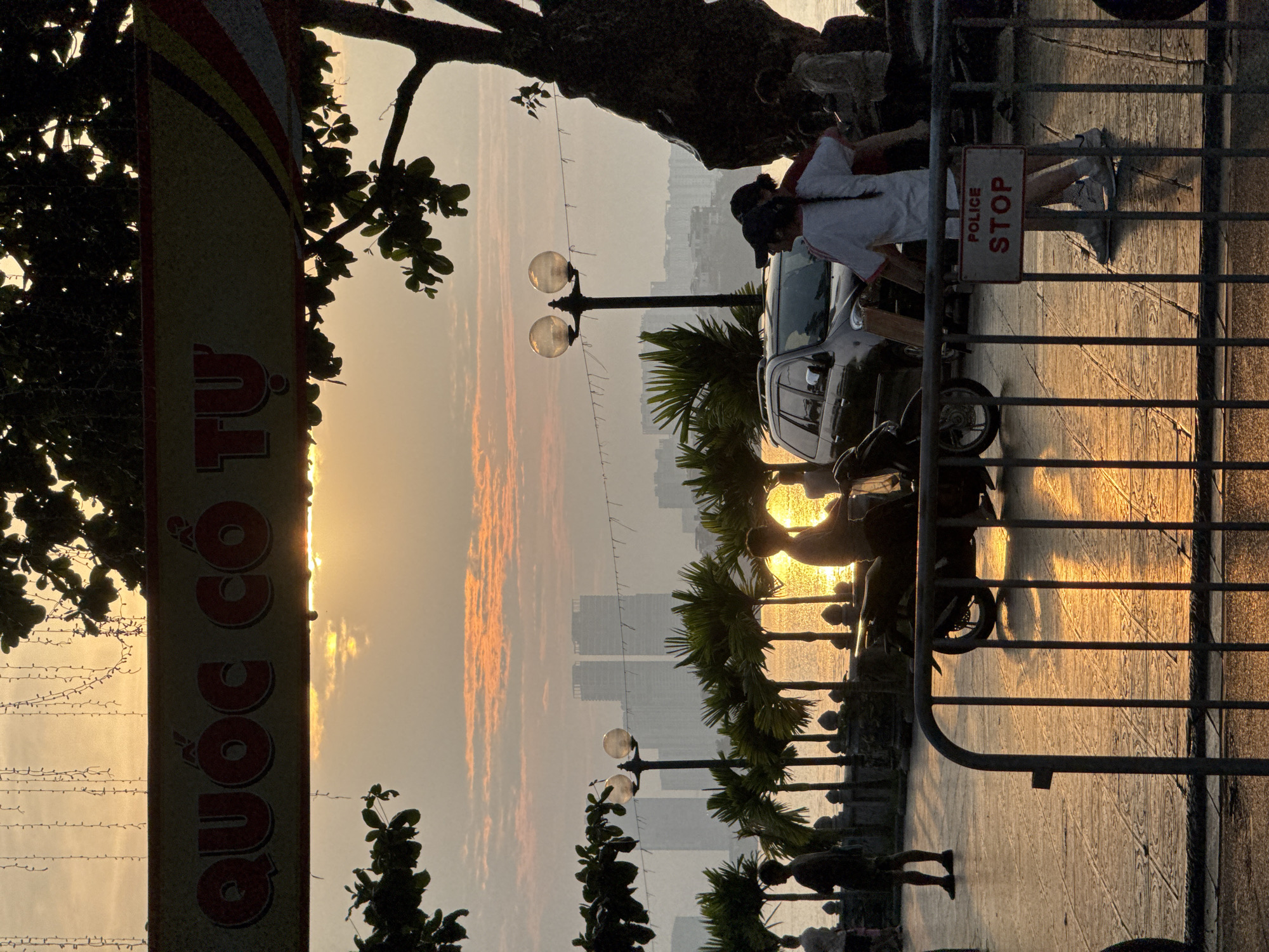 The image depicts a waterfront promenade in Hanoi, Vietnam during a sunset. A banner with Vietnamese text is visible across the top of the frame. In the foreground, a metal barrier with a "POLICE STOP" sign separates the viewer from the scene. Behind the barrier, several individuals are present. A man on a scooter is silhouetted against the water. Two women, one with a braided ponytail, stand near a parked SUV. Other figures are also visible in the background. Palm trees and streetlights line the promenade, and distant buildings are visible across the water. The setting sun casts a warm, golden glow on the water and the wet pavement, creating a serene atmosphere.