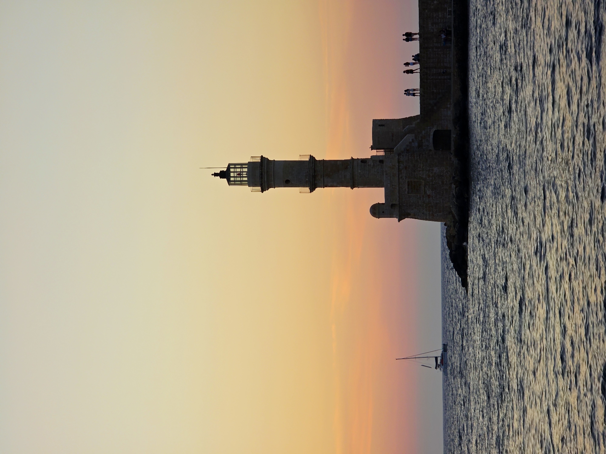 The image depicts the iconic Venetian Lighthouse of Chania, Greece, at sunset. The lighthouse, constructed of stone, stands tall on a breakwater, silhouetted against a sky painted with hues of yellow, orange, and pink. Several small figures, appearing to be people, are visible standing on the breakwater near the lighthouse, observing the sunset or the surroundings. In the sea to the left of the lighthouse, a sailboat with two masts is sailing away from the viewer. The water is dark and choppy, reflecting the warm tones of the sky. The overall scene is serene and picturesque, capturing the beauty of Chania's harbor during twilight.
