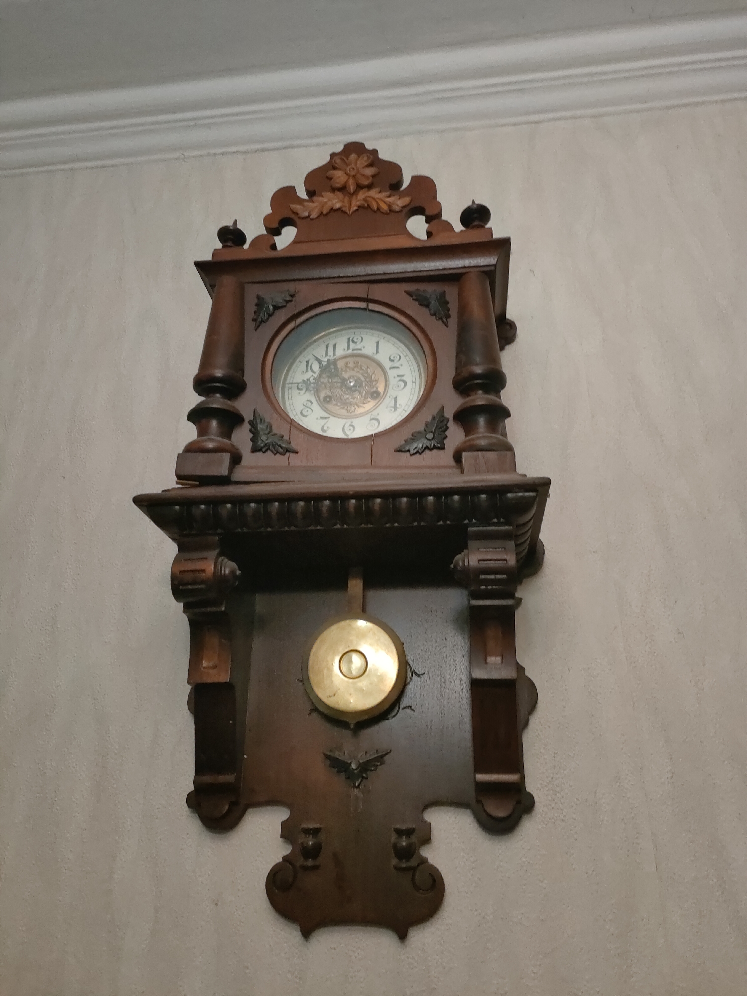 A vertical, eye-level shot shows a dark wood pendulum clock mounted on a textured, light-colored wall. The clock has an ornate top with carved floral and leaf designs, a circular clock face with Roman numerals and decorative hands, and a brass pendulum hanging below. The wall appears to be painted or wallpapered and has a subtle, rough texture. The scene is lit by an unseen light source, casting minimal shadows. This clock is likely located in a residential interior in Minsk, Belarus.