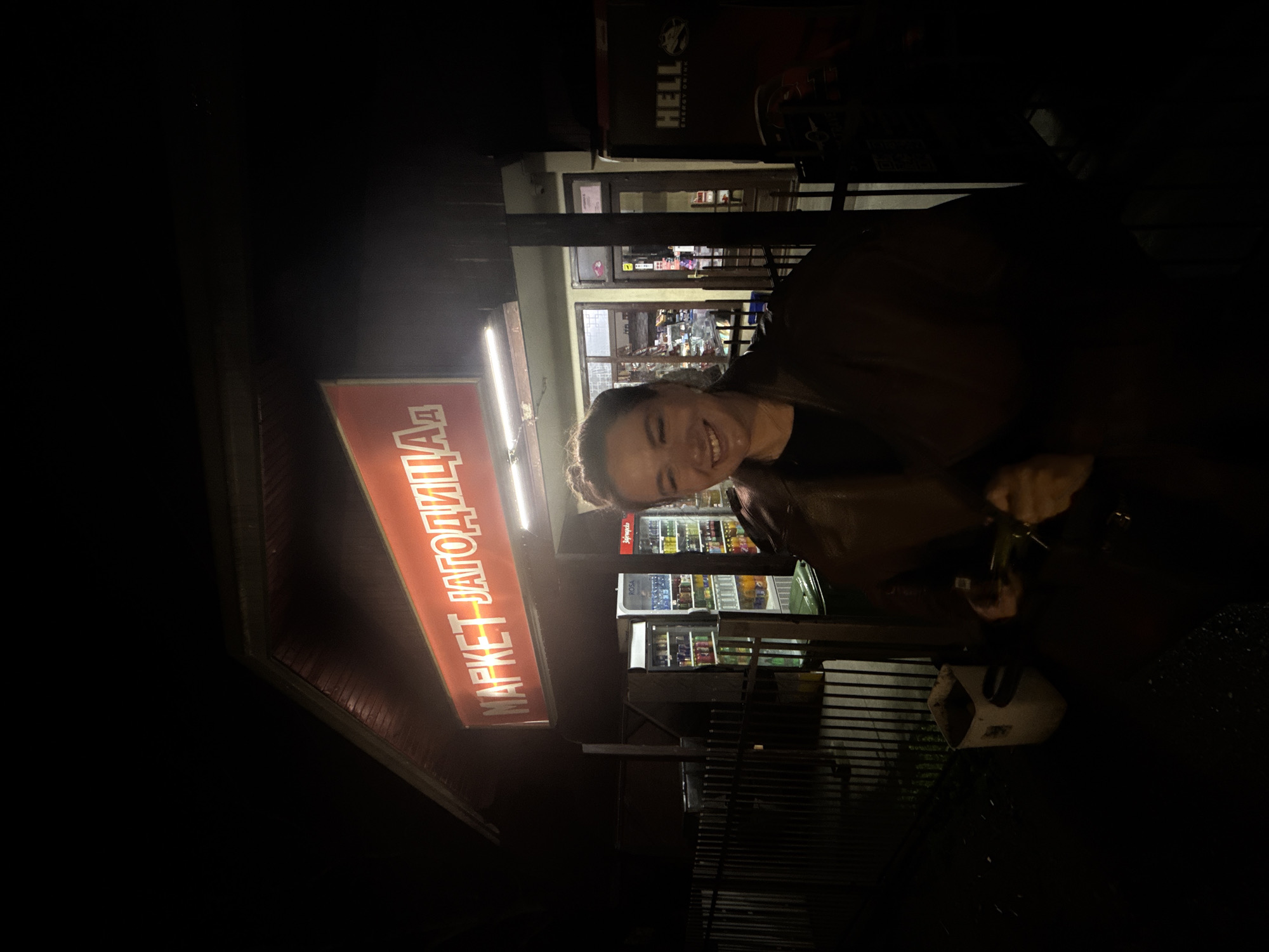 A woman stands outside a small market at night. The red sign reads "MAPKET ЈАГОДИЦА" (MARKET JAGODICA). The market has refrigerated display cases visible through the windows and a "HELL" energy drink advertisement above the entrance. The location is Сремска Рача, Serbia. The woman is smiling and appears to be holding keys. The surrounding environment is dark.