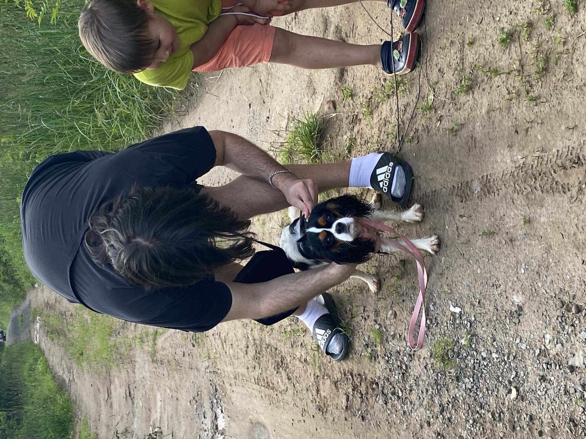 In this image, two individuals and a dog are on a dirt path, likely in a rural or outdoor setting. The primary subject is a person wearing a black t-shirt and black shorts, bent over to interact with a small dog. The dog, a Cavalier King Charles Spaniel, has tri-colored fur and is wearing a pink leash. The person has dark, curly hair and is wearing black Adidas slides with white socks.
To the left and slightly above, a child in a bright lime green t-shirt and orange shorts is visible. The child appears to be looking down, possibly at the dog or the ground. The child is wearing black Crocs with colorful embellishments. The ground is a dirt path with some scattered pebbles and small green plants growing through the soil. To the left of the path, tall green grass is visible.
The overall scene suggests a casual outdoor activity, such as a walk or a break during a hike. The lighting indicates it is daytime, possibly late afternoon or early evening, given the soft light. There are no strong indicators of specific weather, but the presence of lush green grass suggests it's not winter. The setting appears to be a natural environment, possibly a park, trail, or rural road. The interaction between the people and the dog seems relaxed and affectionate.