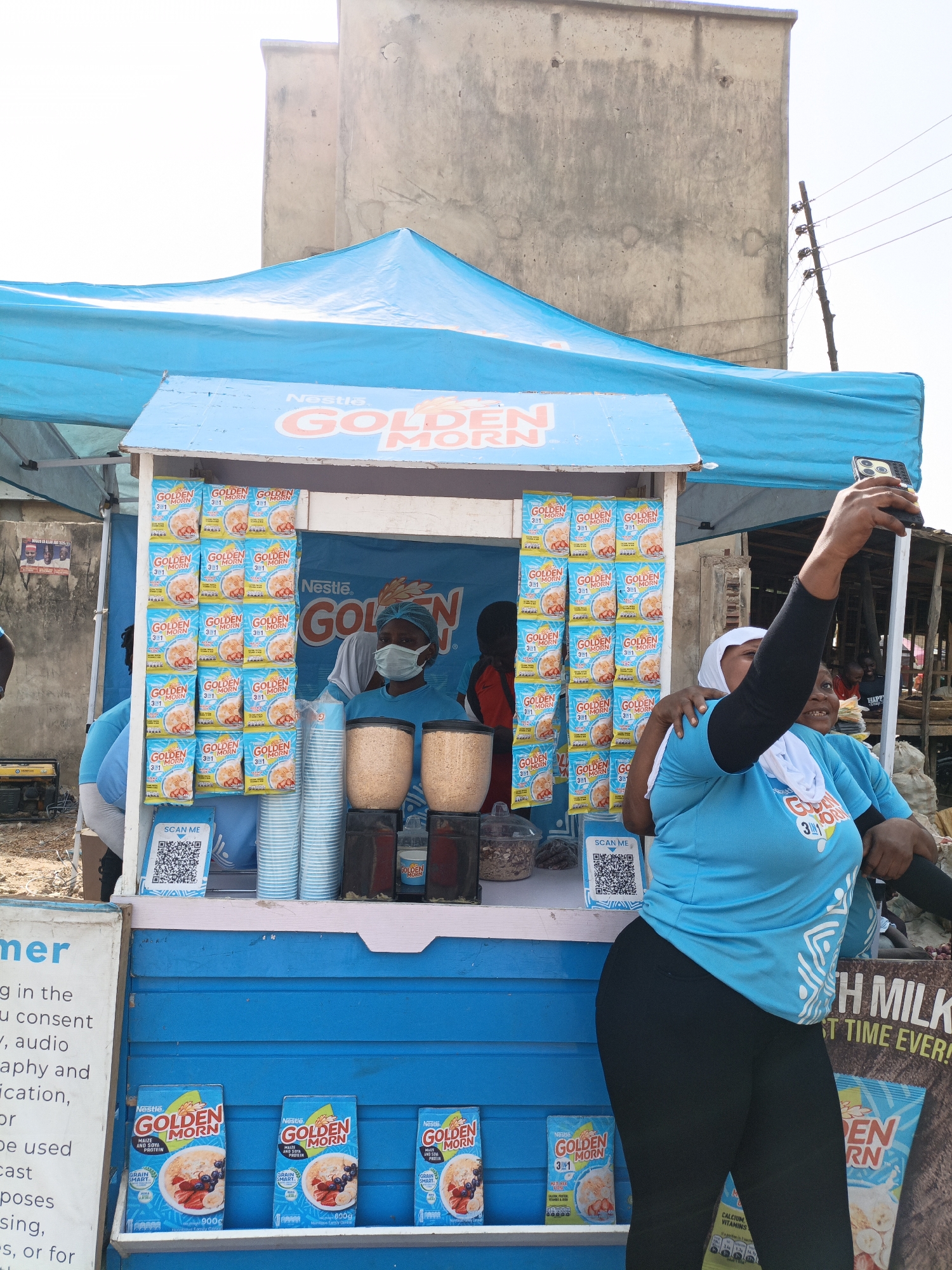 Here's a concise description of the image:
**Location:** Kawaji, Nigeria
**Scene:** An outdoor "Golden Morn" breakfast cereal stand.
**Subjects:**
* A vendor wearing a face mask is standing behind the stand.
* Two women stand on the customer side of the stand, one is holding up a smartphone, presumably taking a selfie. Both are wearing t-shirts branded with "Golden Morn" graphics.
**Objects:**
* A blue-and-white stand with "Nestle Golden Morn" branding, with packages of "Golden Morn" cereal displayed on the shelves.
* Cups and containers are displayed.
* There is additional "Golden Morn" promotional material.
* A blue canopy covers the stand.
* A QR code.
**Actions/Interactions:**
* The woman is holding up a phone, capturing a photograph.
* The vendor is standing behind the stand, presumably selling the product.
**Environment:** A busy outdoor market setting. A building is visible in the background.