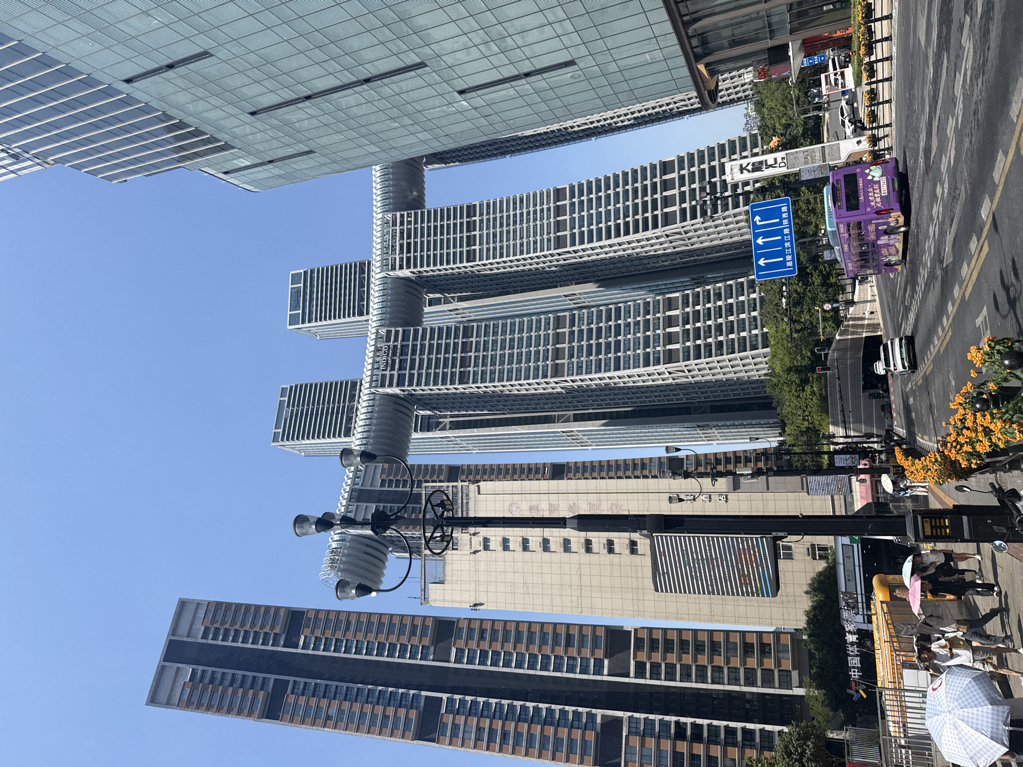 The image shows a street scene in Yuzhong District, China, with several tall buildings dominating the skyline. In the foreground, a street is visible with a purple bus and several cars driving on it. Pedestrians are walking on the sidewalk, some holding umbrellas to shield themselves from the sun. A street lamp with multiple lights stands prominently in the mid-ground. The background features a cluster of modern skyscrapers, including the iconic Raffles City Chongqing, characterized by its unique "sail" or "ship" design connecting multiple towers. The sky is clear and blue. The architecture suggests a bustling urban environment.