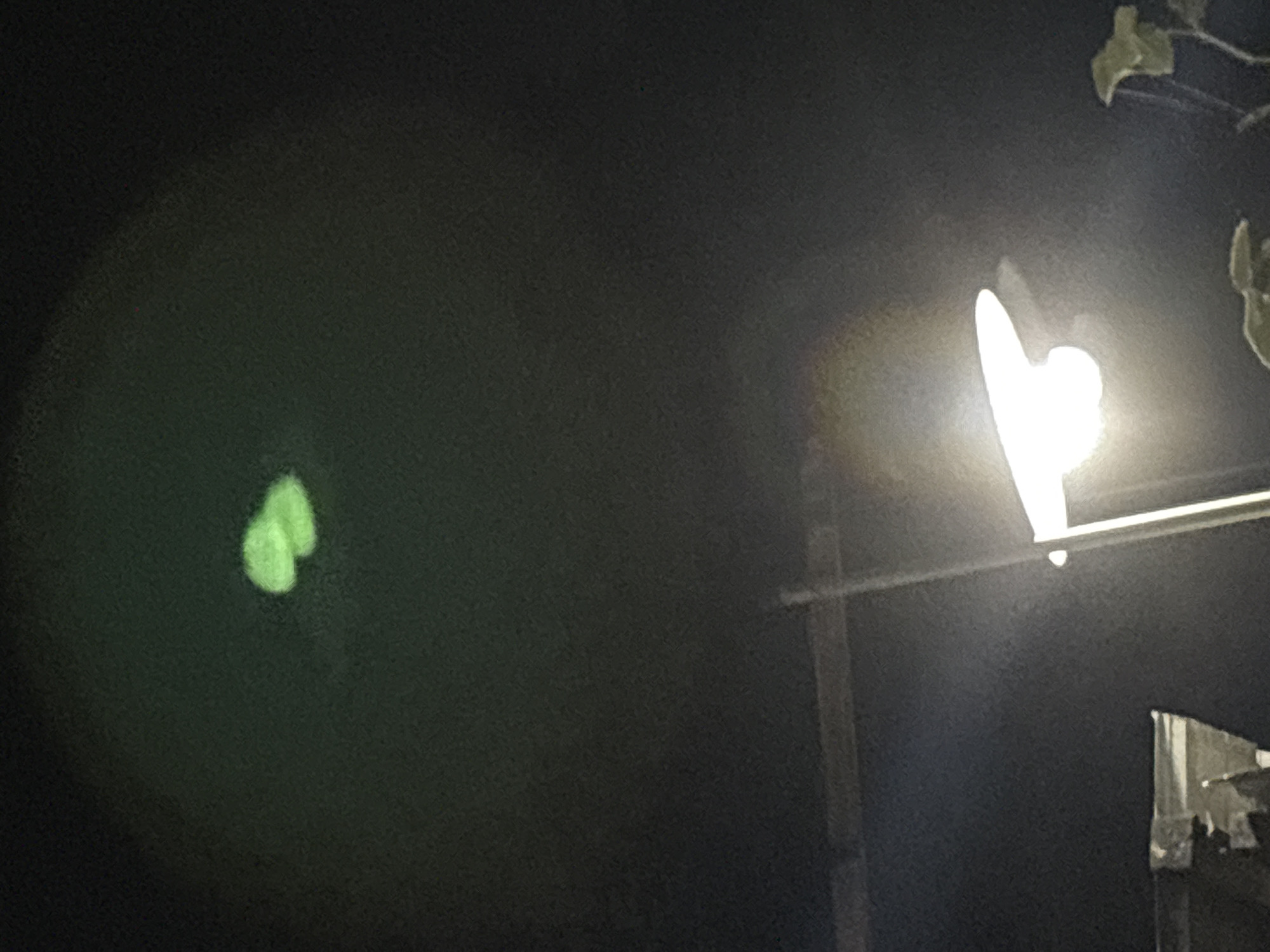 In the dark, at night, a bright street lamp illuminates the scene. Above the lamp, in the upper portion of the image, a blurry, faint, greenish object is visible. The object appears to be amorphous in shape, with indistinct edges. The background is dark, with some faint structures visible at the bottom of the frame, suggesting an outdoor environment in an urban or suburban setting. The image's focus is on the street lamp and the unexplained aerial object. The location is identified as Gempol, Indonesia.