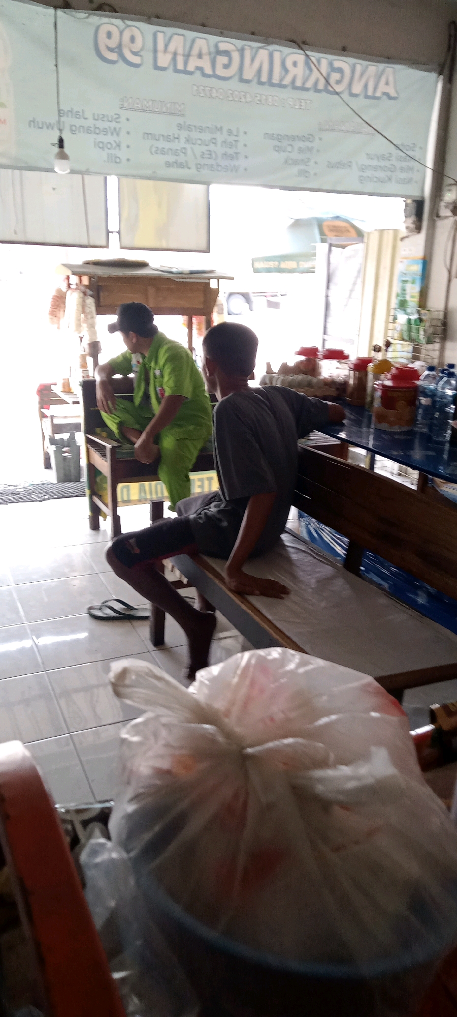 The image depicts an outdoor food stall in Delanggu, Indonesia. The stall has a banner overhead displaying prices, with text like "ANEKA MINUMAN" (assorted drinks) and "MENU" (menu). Below the banner, a menu list includes items such as "Es Mineral", "Teh Pucuk", "Kopi", and "Jus". Two men are seated at the stall. One man, wearing a green uniform and a black cap, is sitting on a wooden bench. The second man, wearing a grey shirt, is also seated on a bench. In the foreground, a plastic bag filled with goods obscures the view of other items. The environment appears to be a street or roadside location, indicated by the tiled floor and the open-air setup of the stall.