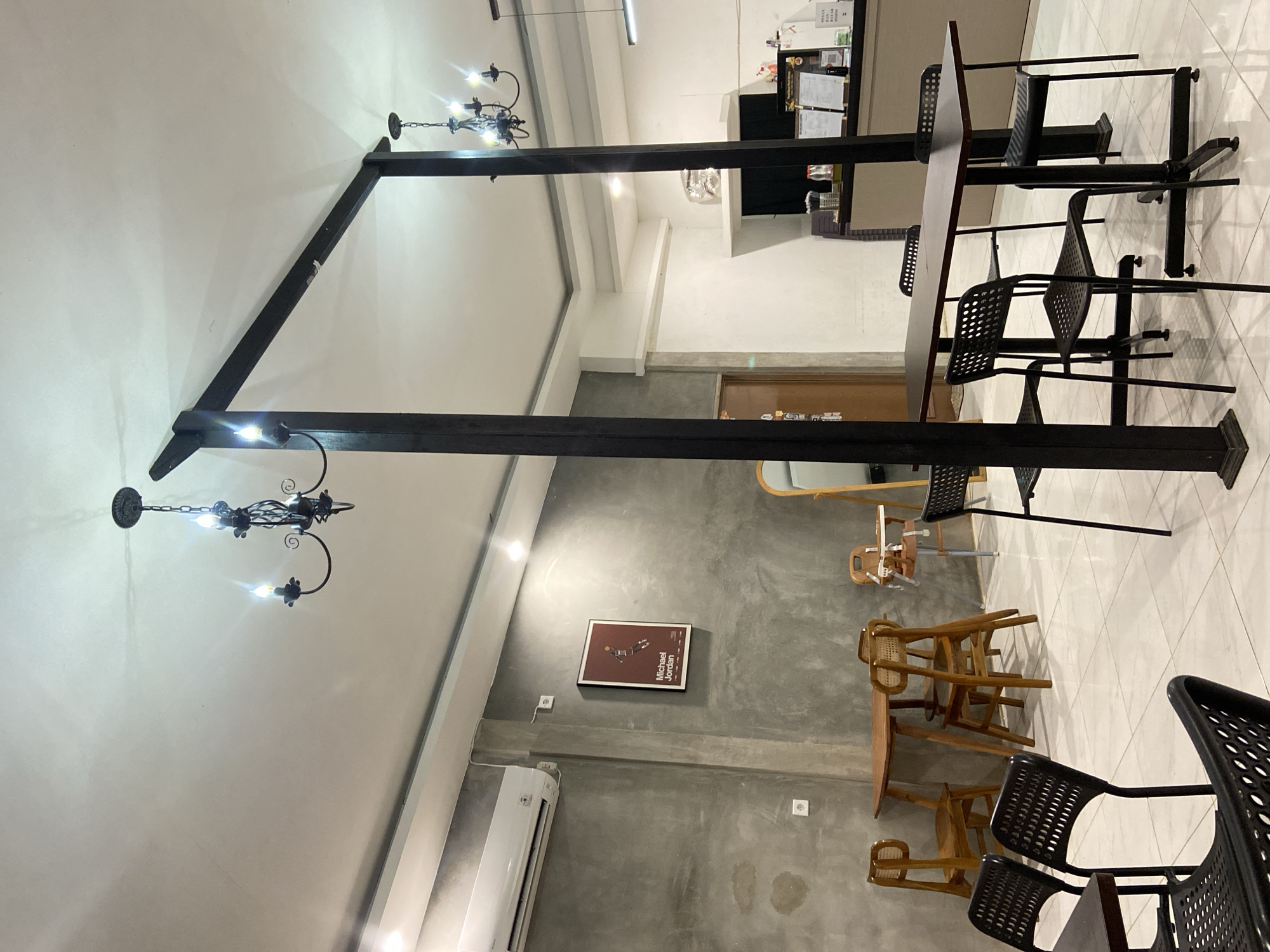 This media file depicts the interior of a modern cafe or restaurant. The scene is illuminated by artificial lighting, suggesting it could be any time of day. The setting is characterized by a blend of industrial and natural elements. A prominent feature is a large, dark, angled metal beam structure, possibly a decorative element or part of the building's architecture, which cuts across the frame.
Several tables and chairs are arranged throughout the space. There are dark, perforated metal chairs around dark-topped tables, suggesting a contemporary seating area. In another section, there are several wooden chairs with woven seats, some stacked and others arranged around wooden tables, hinting at a more rustic or casual dining area. One of these wooden tables has a chair with a high back and tray, possibly a high chair for a child.
On a grey concrete wall, there is a framed poster with an image and the text "Michael Jordan". Below this poster, a white air conditioning unit is mounted. Two wall outlets are also visible on the concrete wall.
The floor is tiled with a light-colored square pattern. A door with a wooden frame is visible in the background on the right side of the image. The overall ambiance appears calm and inviting, typical of a contemporary establishment. No people are visible in the frame. The location context provided is Sekar Mawar, Indonesia, which might suggest the architectural style or decor could be influenced by local aesthetics, though this is not explicitly evident from the visual elements alone.